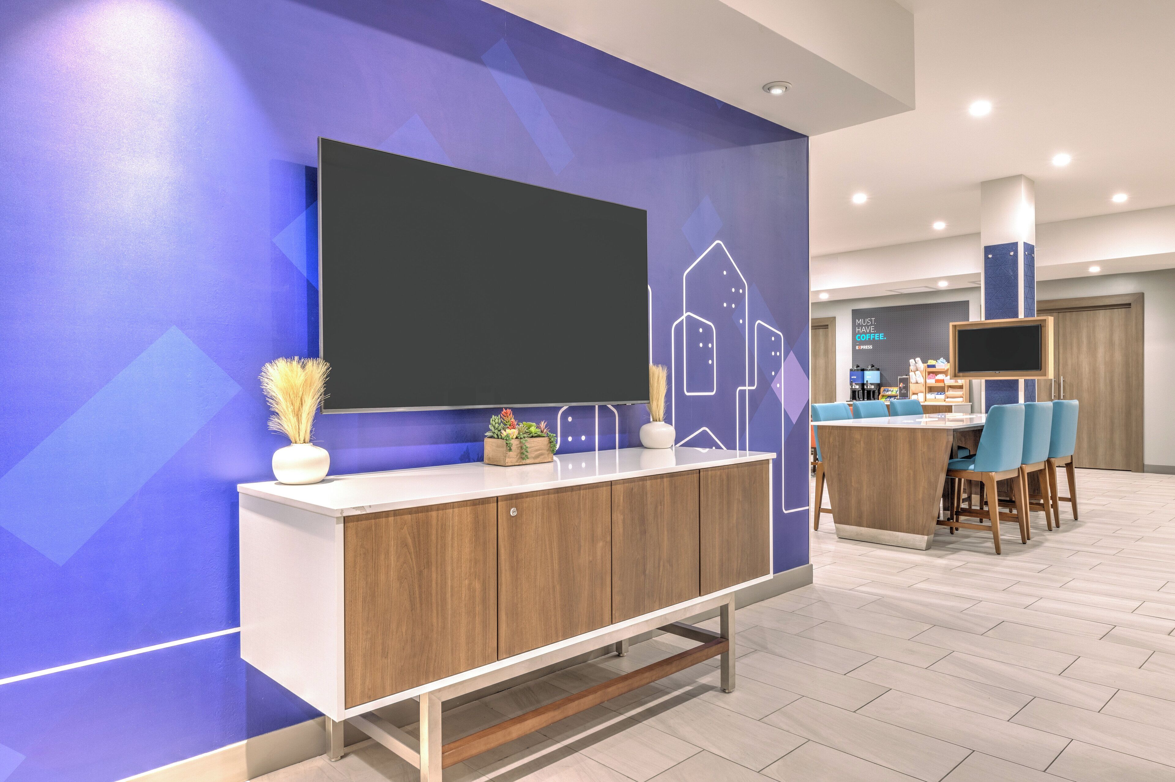 Una moderna hall d'albergo con una grande televisione a schermo piatto montata a parete, una reception con un mobile in legno e un'area relax con poltrone blu.