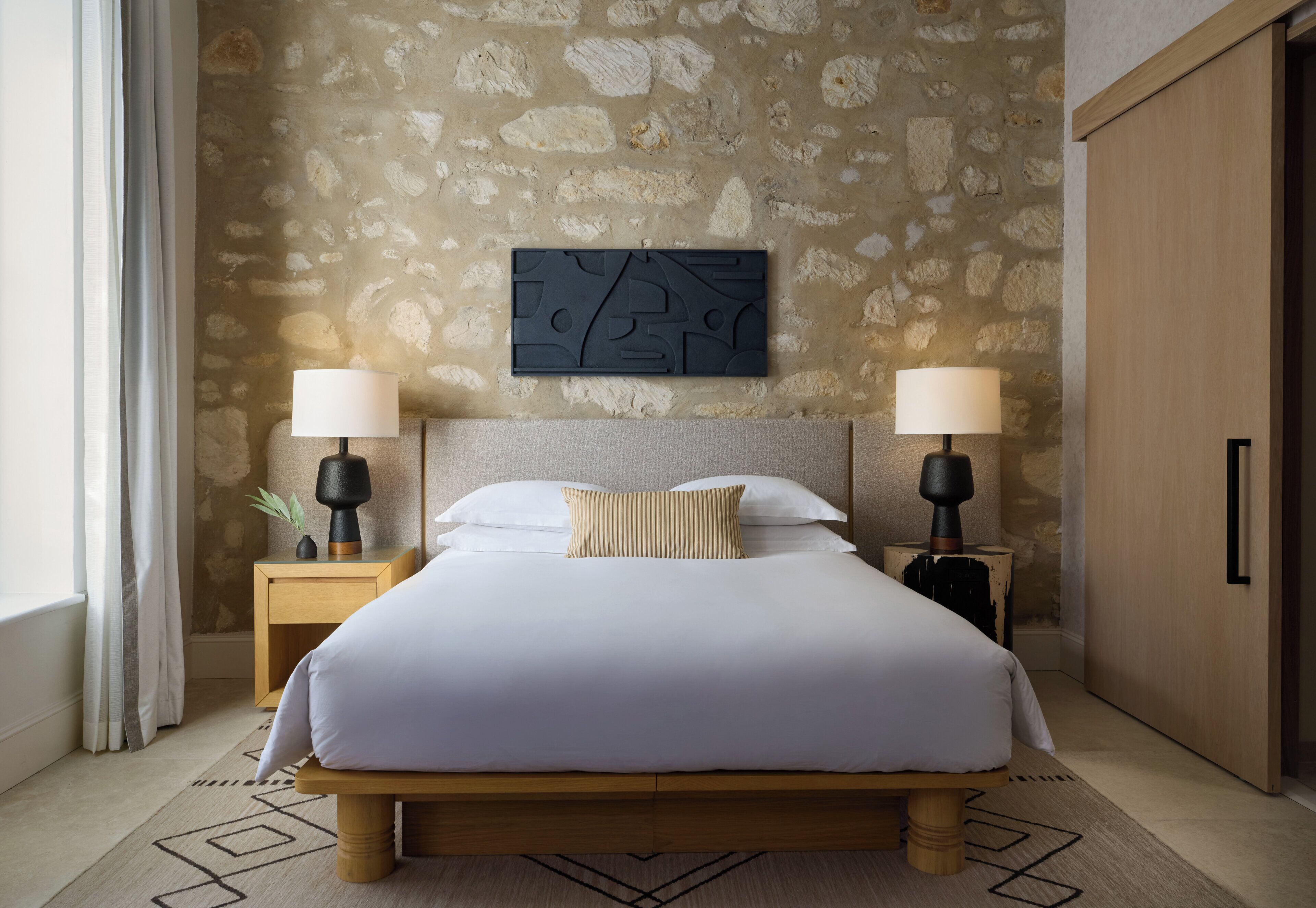 A neatly made bed with white bedding and a striped pillow, flanked by two bedside tables with lamps, set against a stone wall with a geometric artwork above.