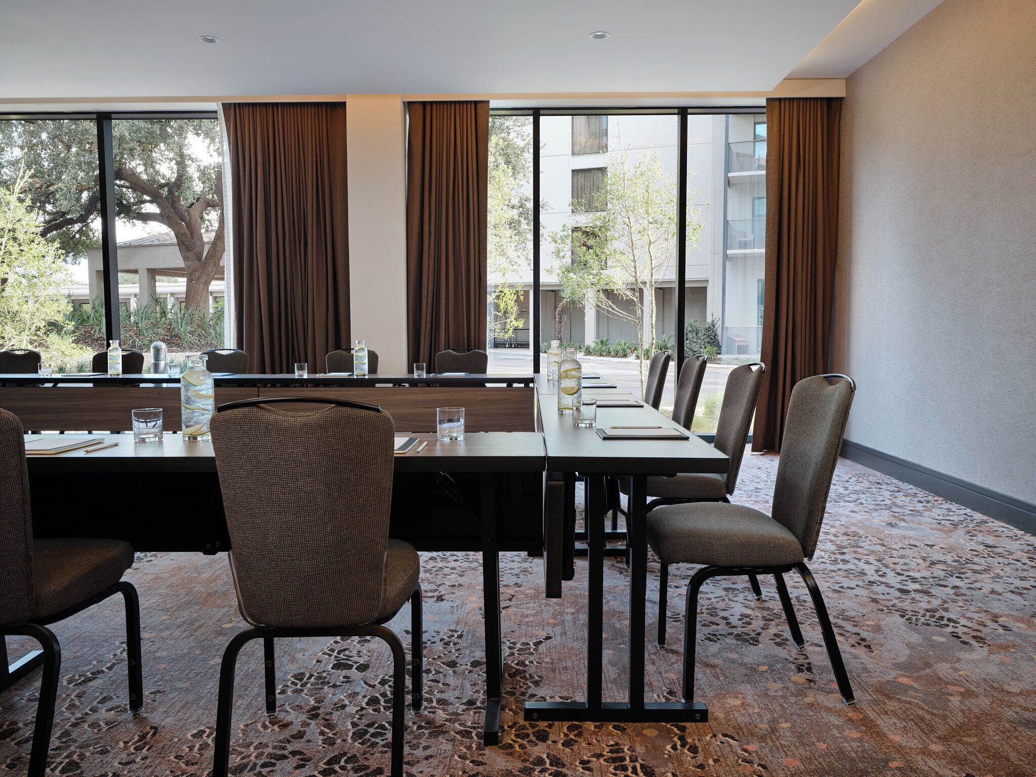 Un salón de conferencias con una larga mesa, sillas y vistas a un edificio y árboles a través de amplios ventanales.