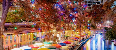 A riverside walkway with outdoor seating under umbrellas, trees adorned with lights, and a building in the background.