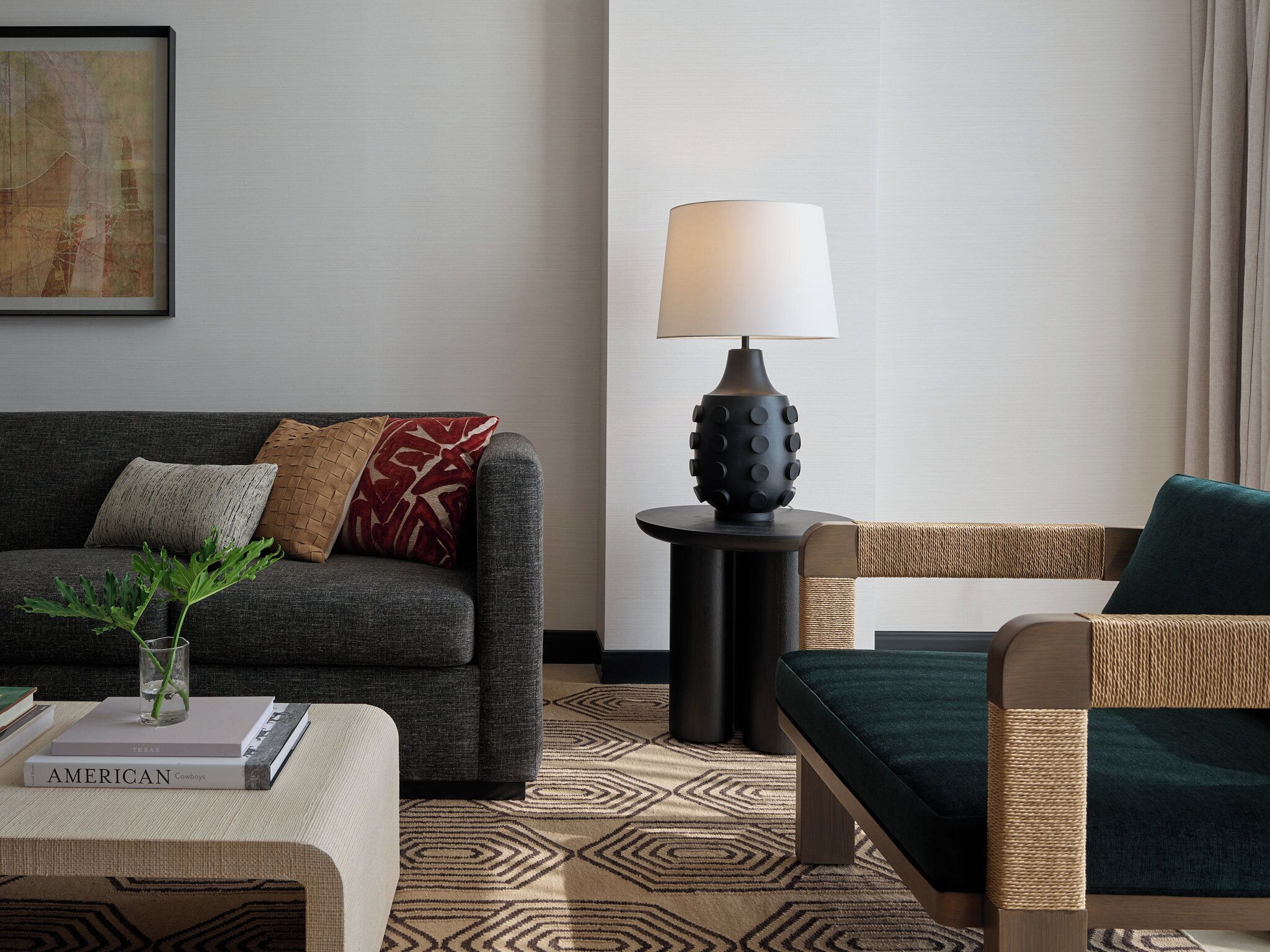 Ein modernes Wohnzimmer mit einem grauen Sofa, einem runden Beistelltisch mit Lampe, einem Korbstuhl und einem gemusterten Teppich.