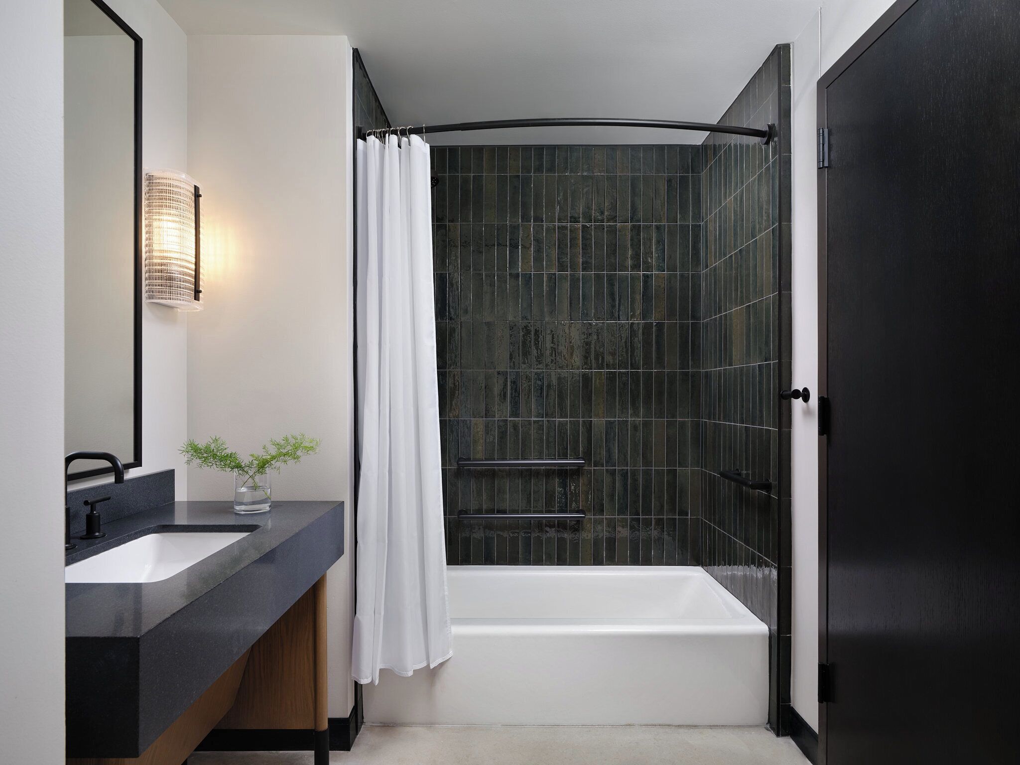 A modern bathroom with a dark tiled shower, a white bathtub, and a black countertop with a mirror and a small plant.