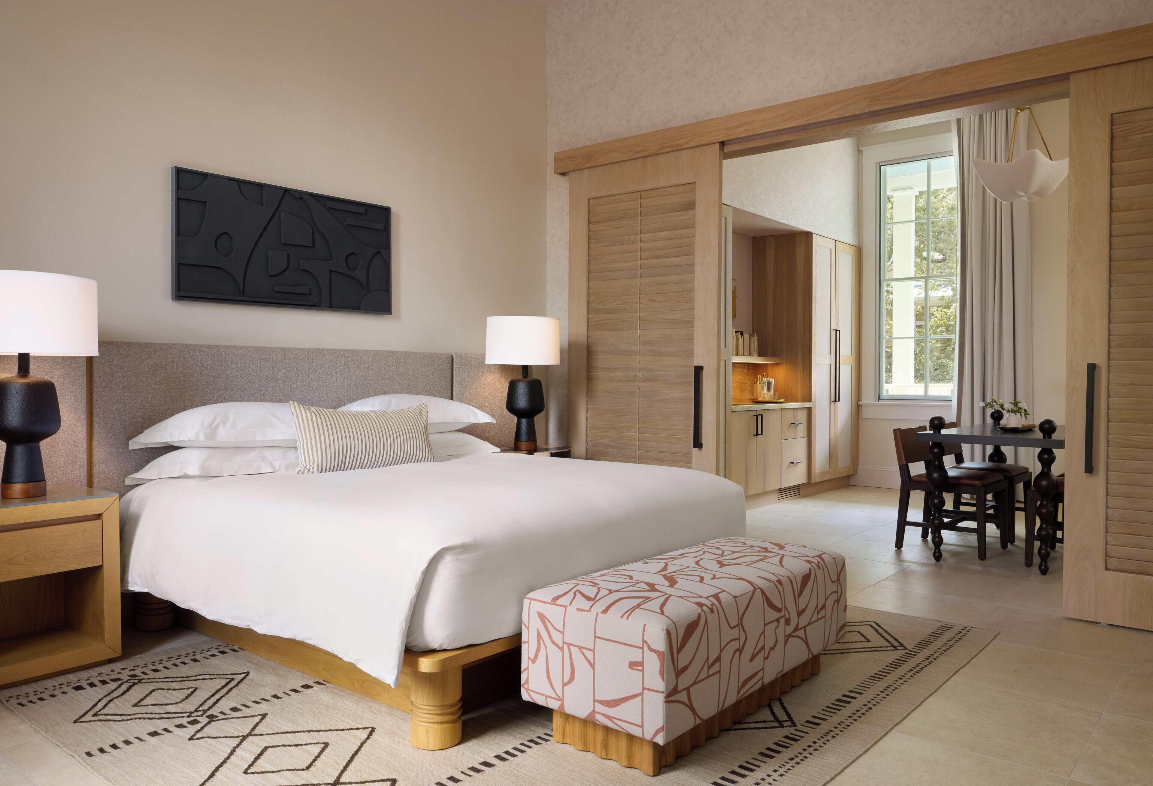 Una habitación de hotel moderna con una cama grande, lámparas de noche, un escritorio con una silla y vistas al exterior a través de una puerta corredera.