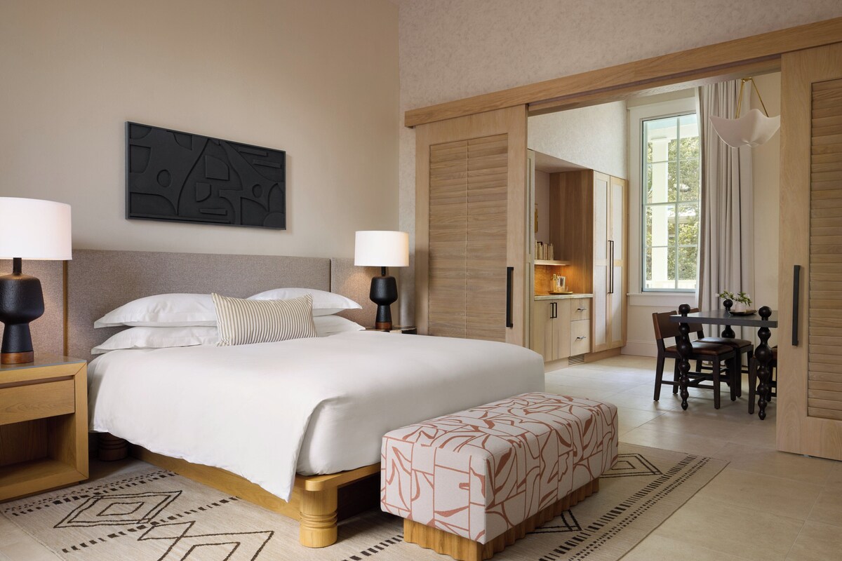 A modern hotel room with a large bed, bedside lamps, a desk with a chair, and a view of the outdoors through a sliding door.