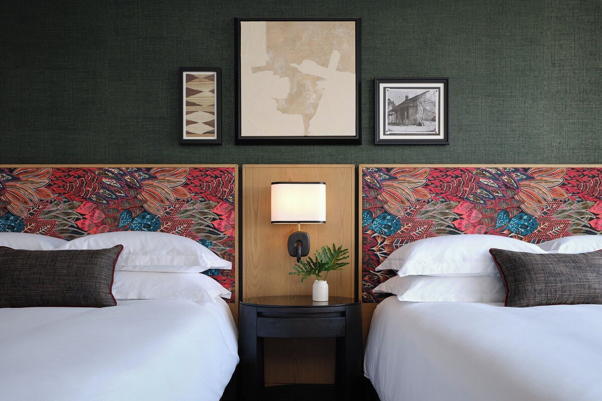 A hotel room with two beds, a nightstand with a lamp, and a wall with three framed artworks.