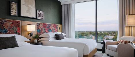 Habitación de hotel con dos camas, un ventanal y vista a la ciudad, una silla y una lámpara.