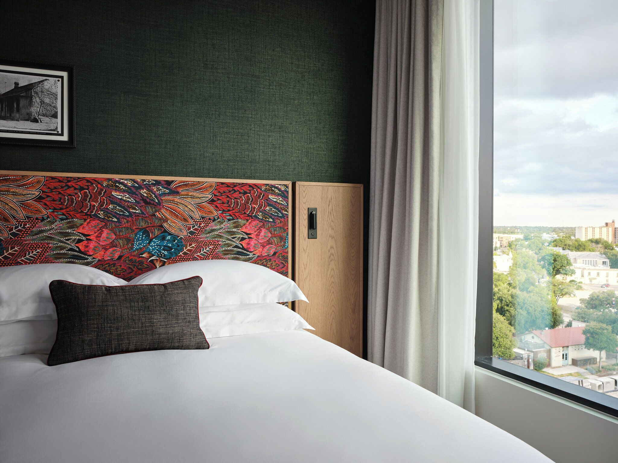 A hotel room with a bed, a headboard with a colorful pattern, a window with a view of buildings, and a framed picture on the wall.