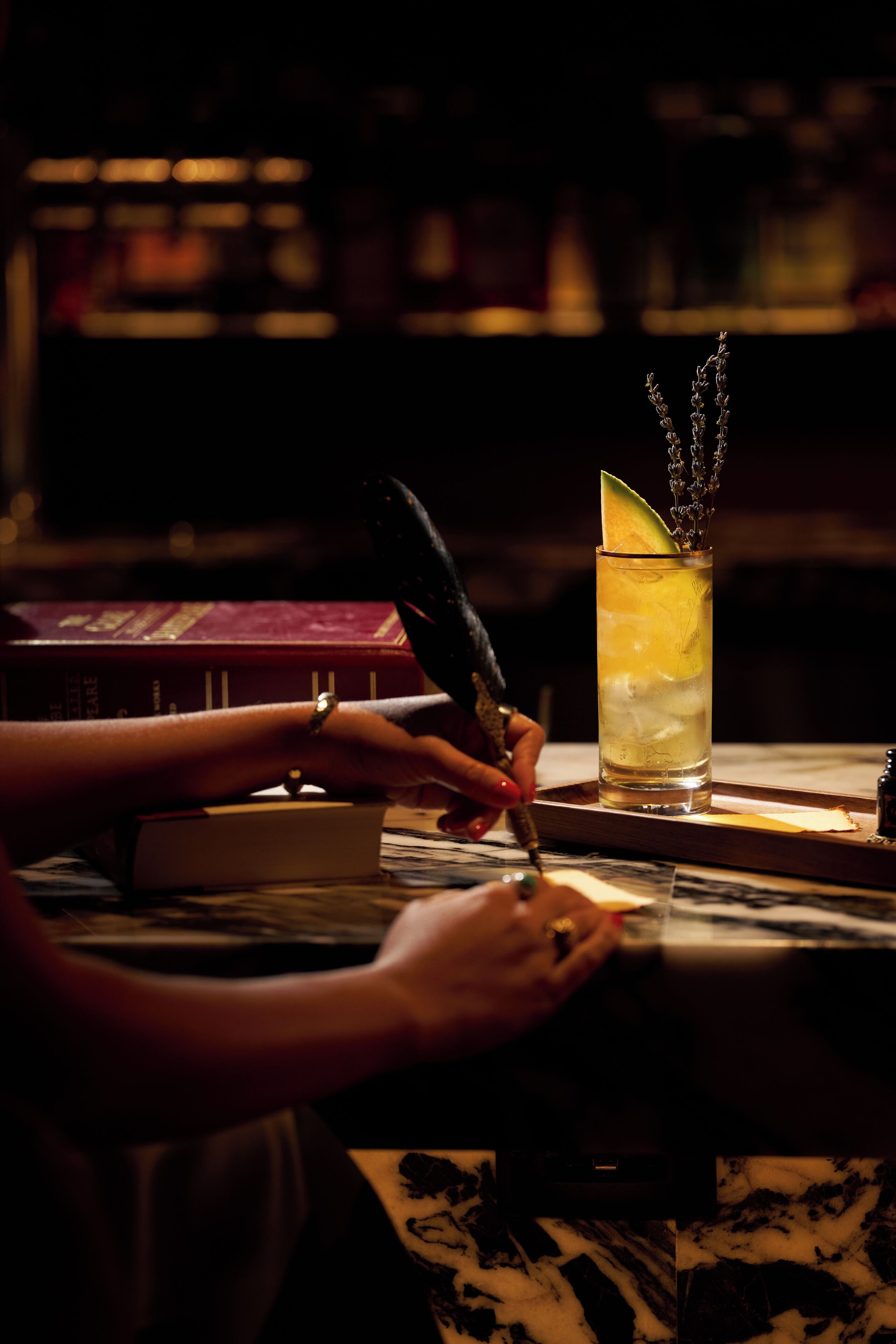 A person writing with a pen on a table with a glass of drink and a book in the background.