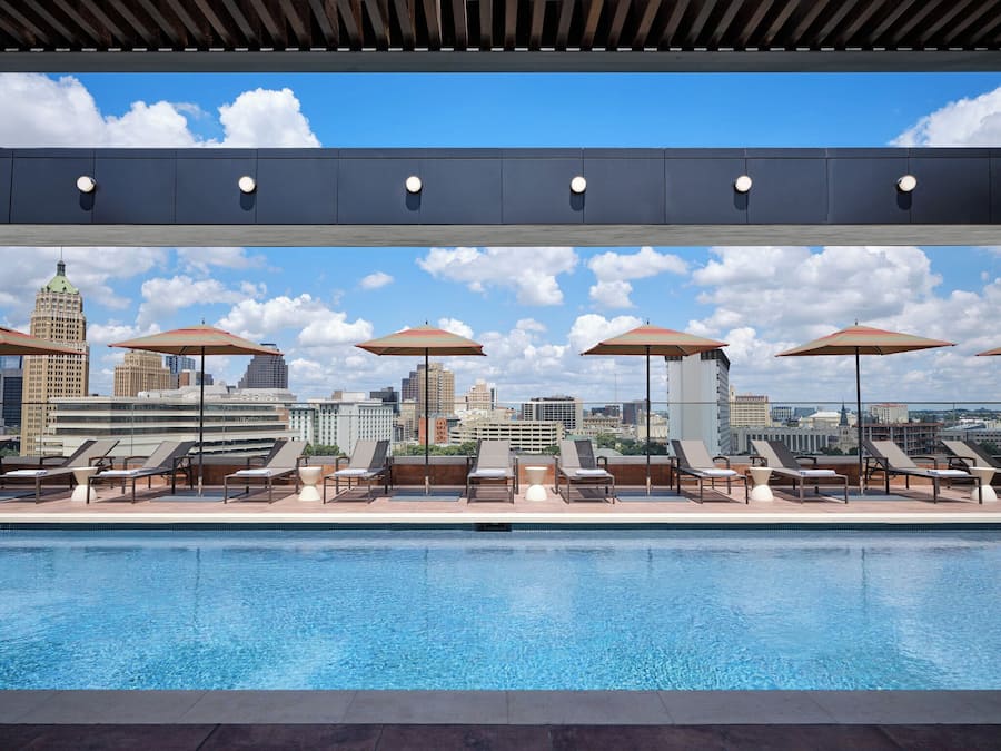 A rooftop pool with lounge chairs and umbrellas, overlooking a city skyline.