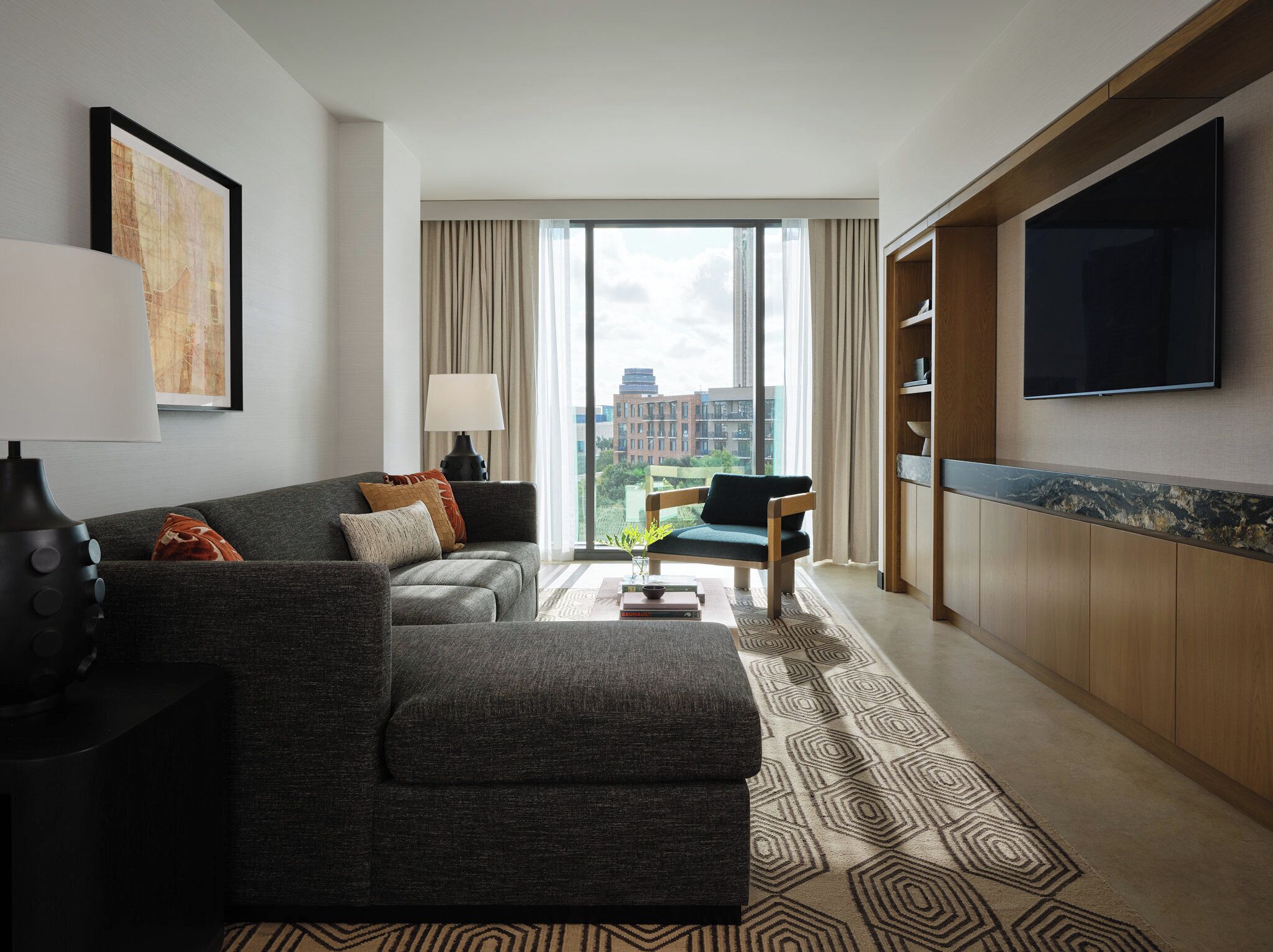 Une chambre d’hôtel moderne avec un canapé gris, une table d’appoint en bois, un téléviseur à écran plat et une grande fenêtre donnant sur la ville.