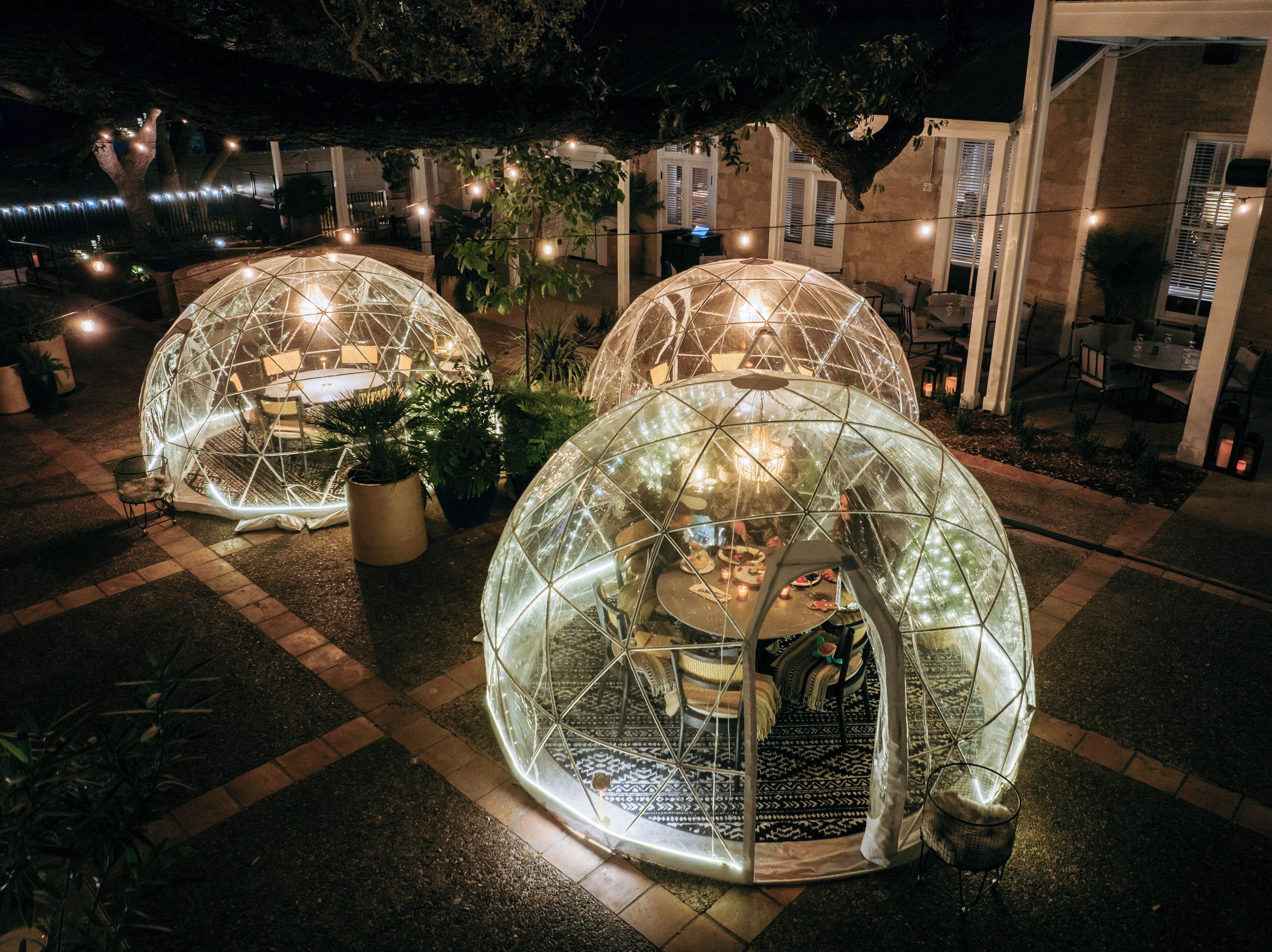 Deux structures en forme de dôme géodésique transparentes, avec des tables et des chaises à l’intérieur, entourées de plantes en pot et illuminées par des guirlandes lumineuses.