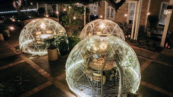 Dos estructuras geodésicas transparentes con mesas y sillas en su interior, rodeadas de plantas en macetas y luces colgantes.