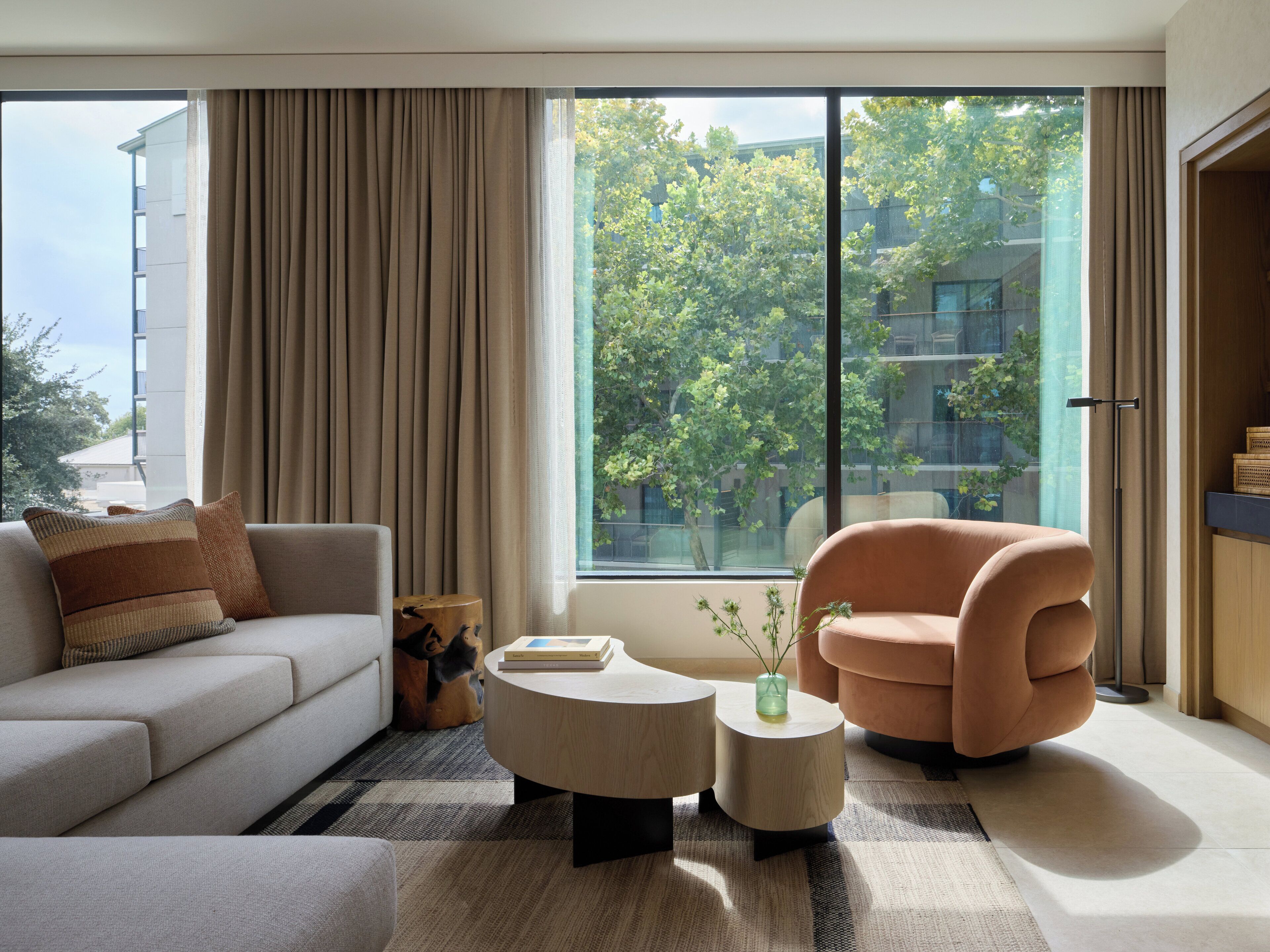 A modern living room with a sofa, armchair, and coffee table, large windows with a view of trees and buildings, and a floor lamp.