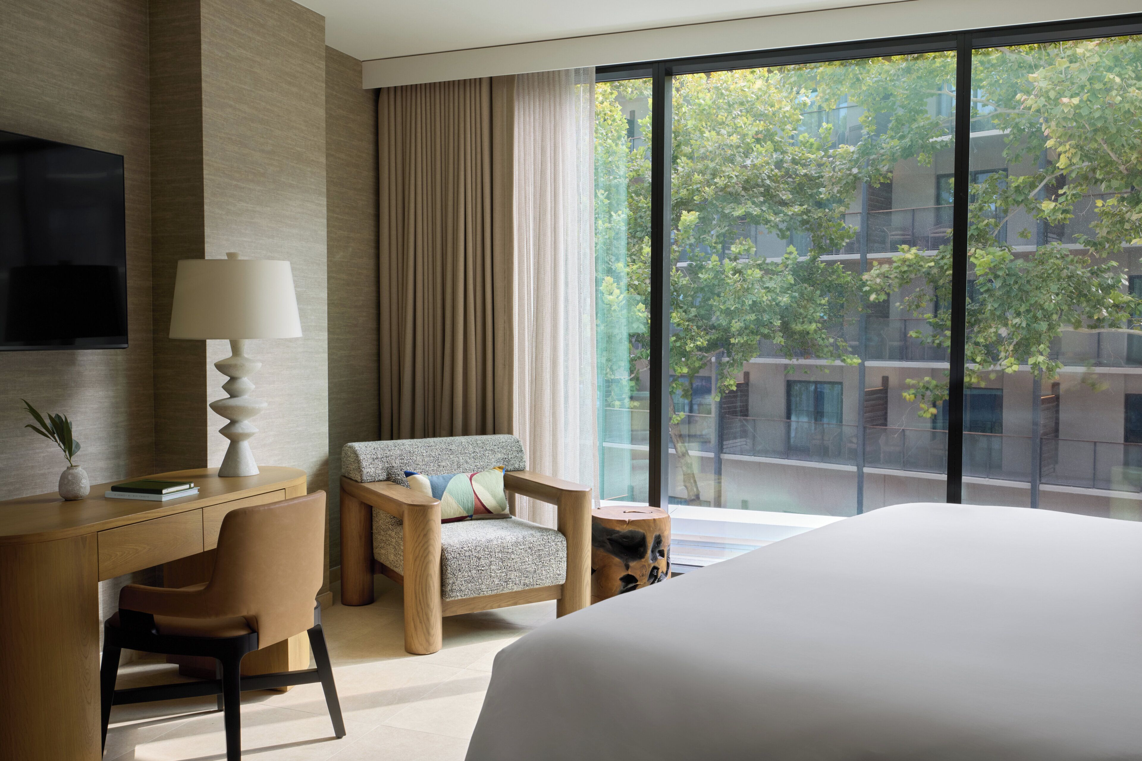 Una habitación de hotel moderna con una cama grande, un escritorio con lámpara, una silla y vistas a un paisaje urbano a través de amplios ventanales.