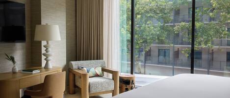 A modern hotel room with a large bed, a desk with a lamp, a chair, and a view of a cityscape through large windows.