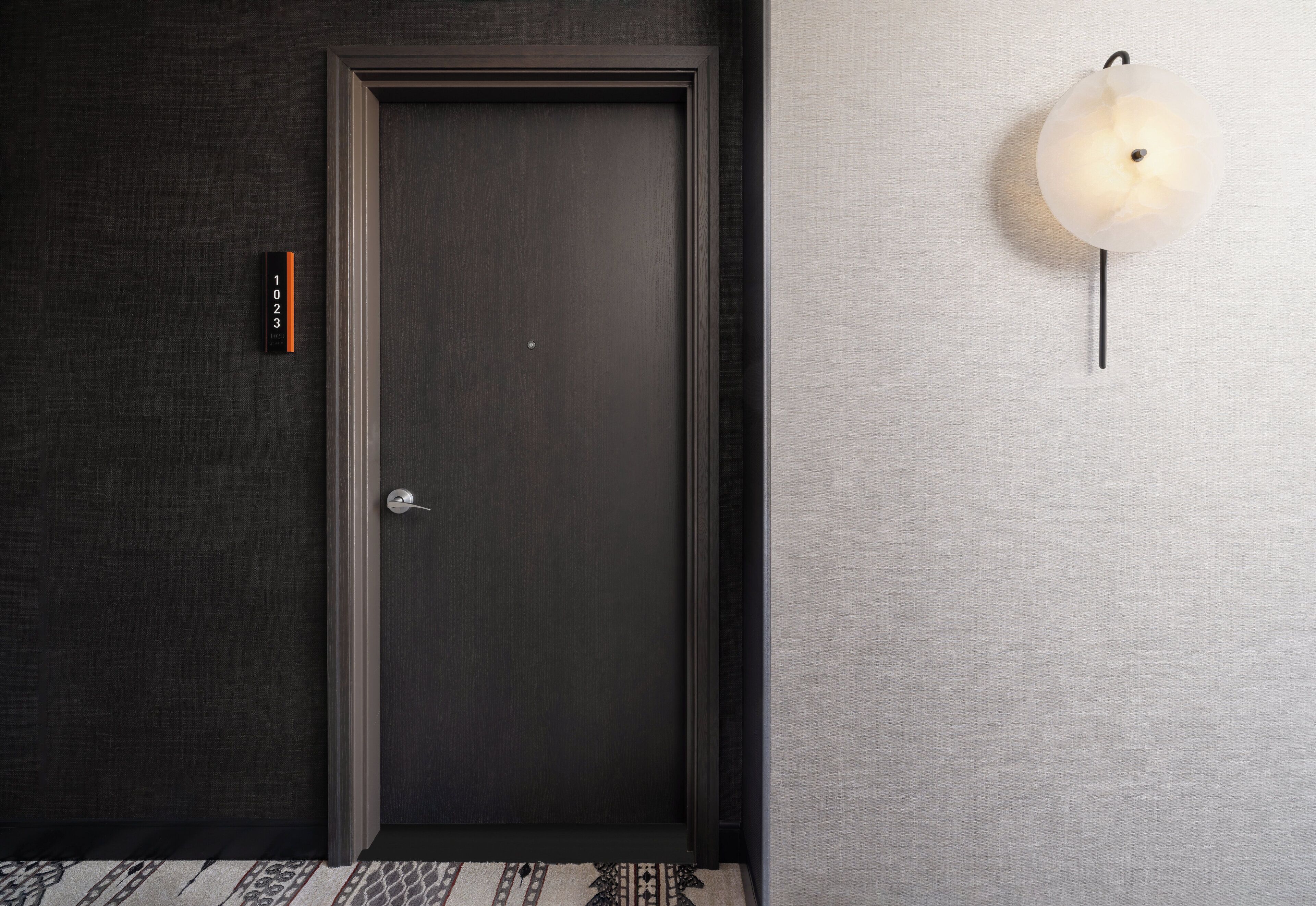 A dark wooden door with a silver handle, a small sign with numbers, and a wall-mounted light fixture.