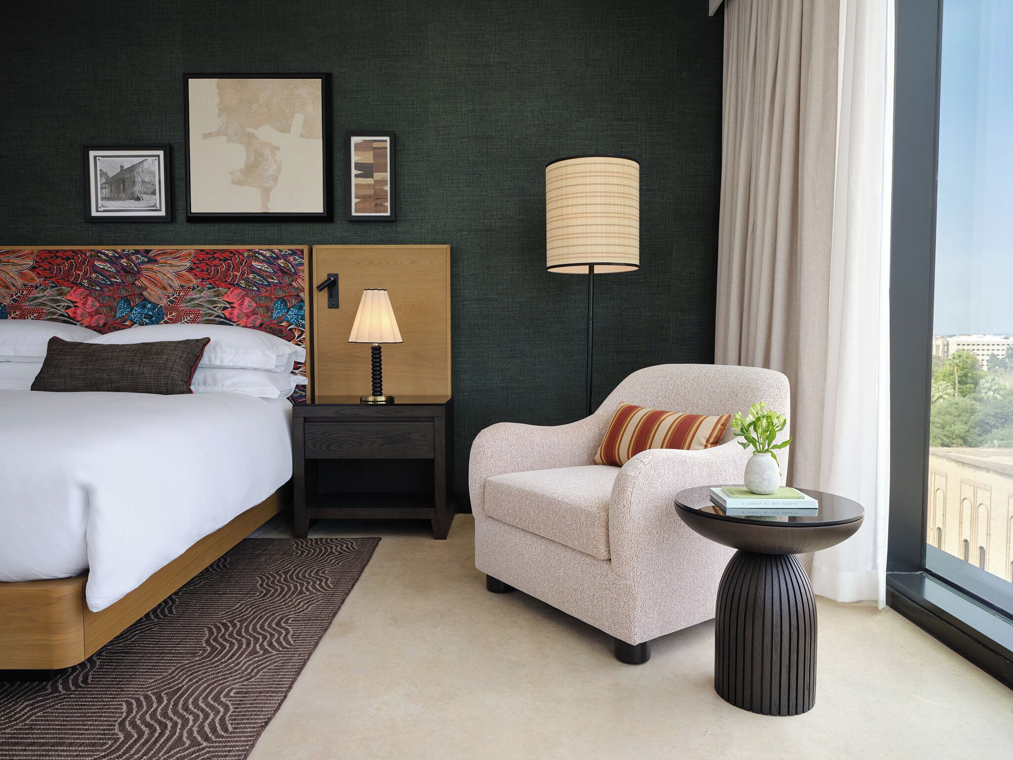 A hotel room with a bed, a nightstand with a lamp, a chair, a small table with a plant, and a view of the city through the window.