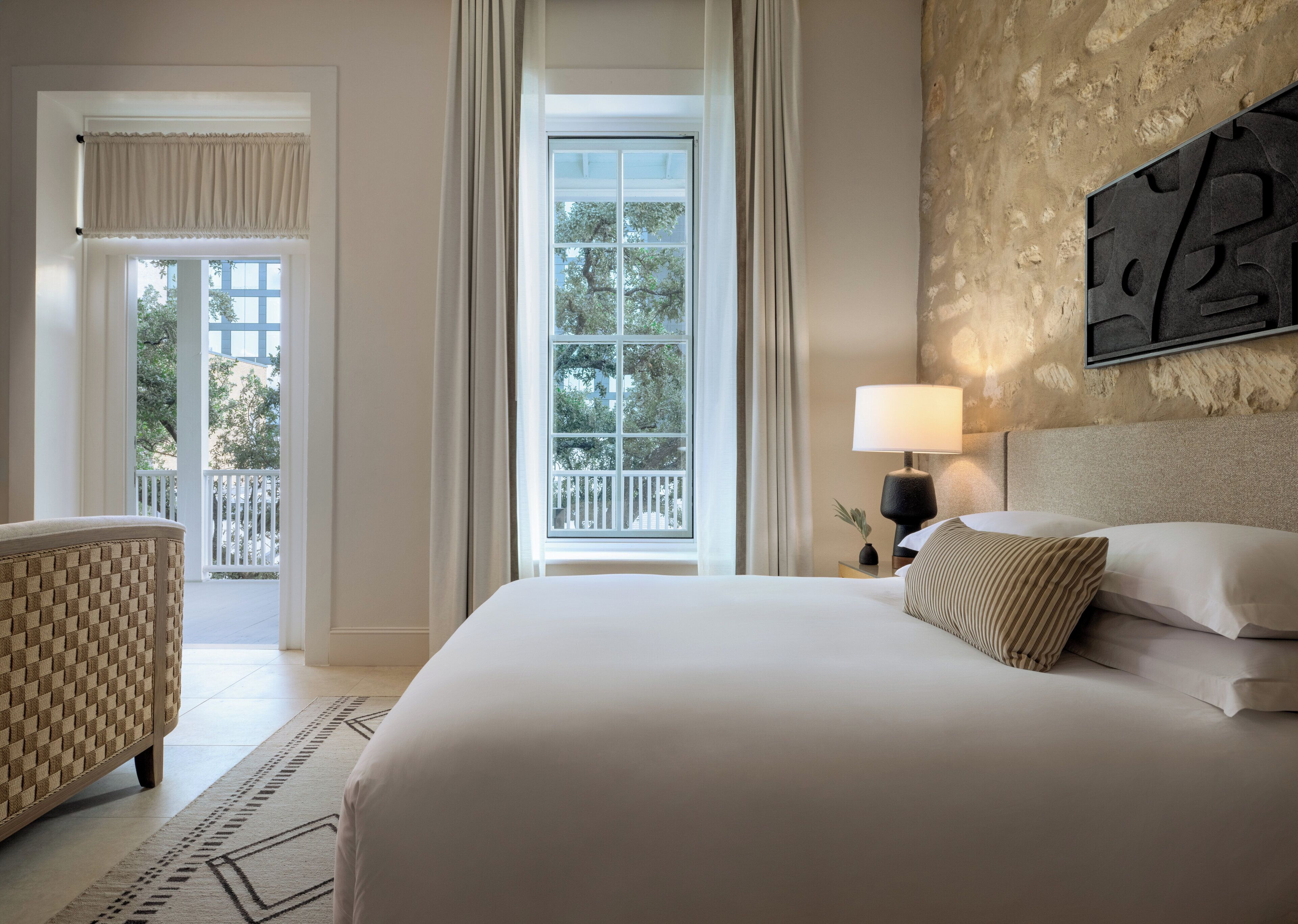 Une chambre d’hôtel avec un grand lit, une table de chevet avec une lampe, une vue sur l’extérieur par une fenêtre et un balcon.