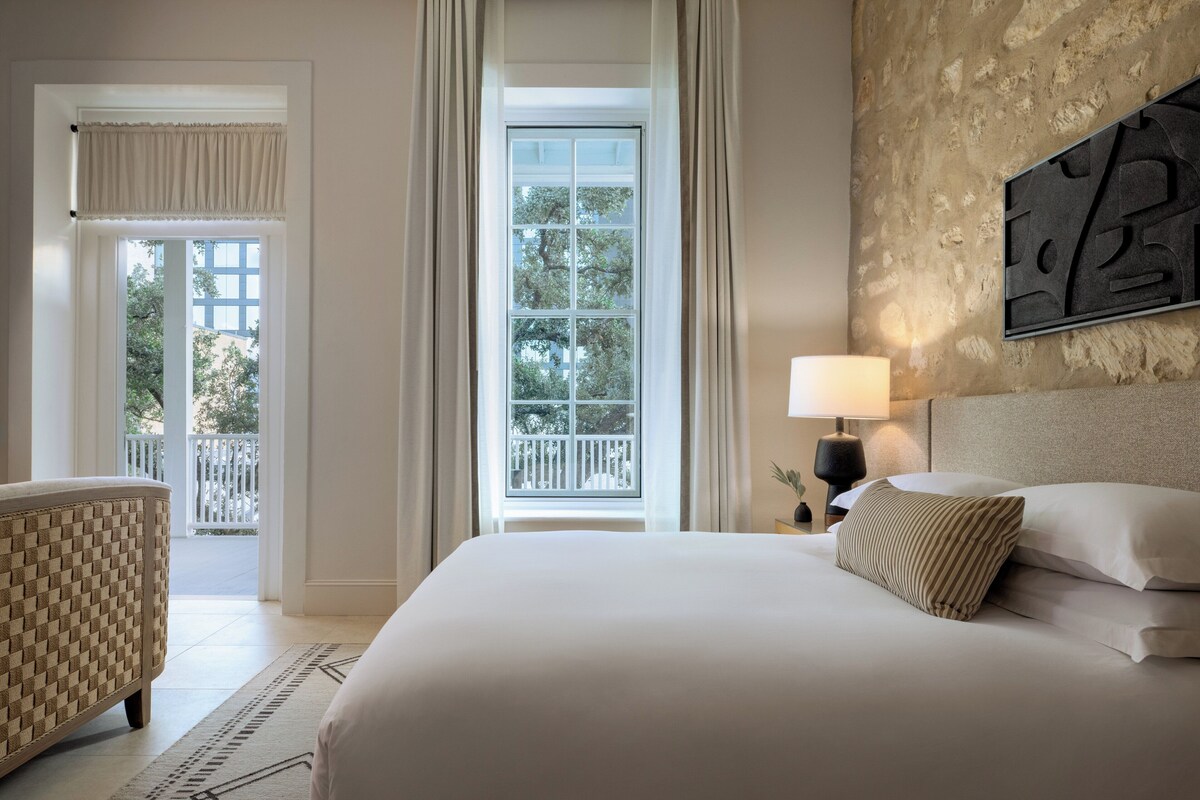 A hotel room with a large bed, a nightstand with a lamp, a view of the outdoors through a window, and a balcony.