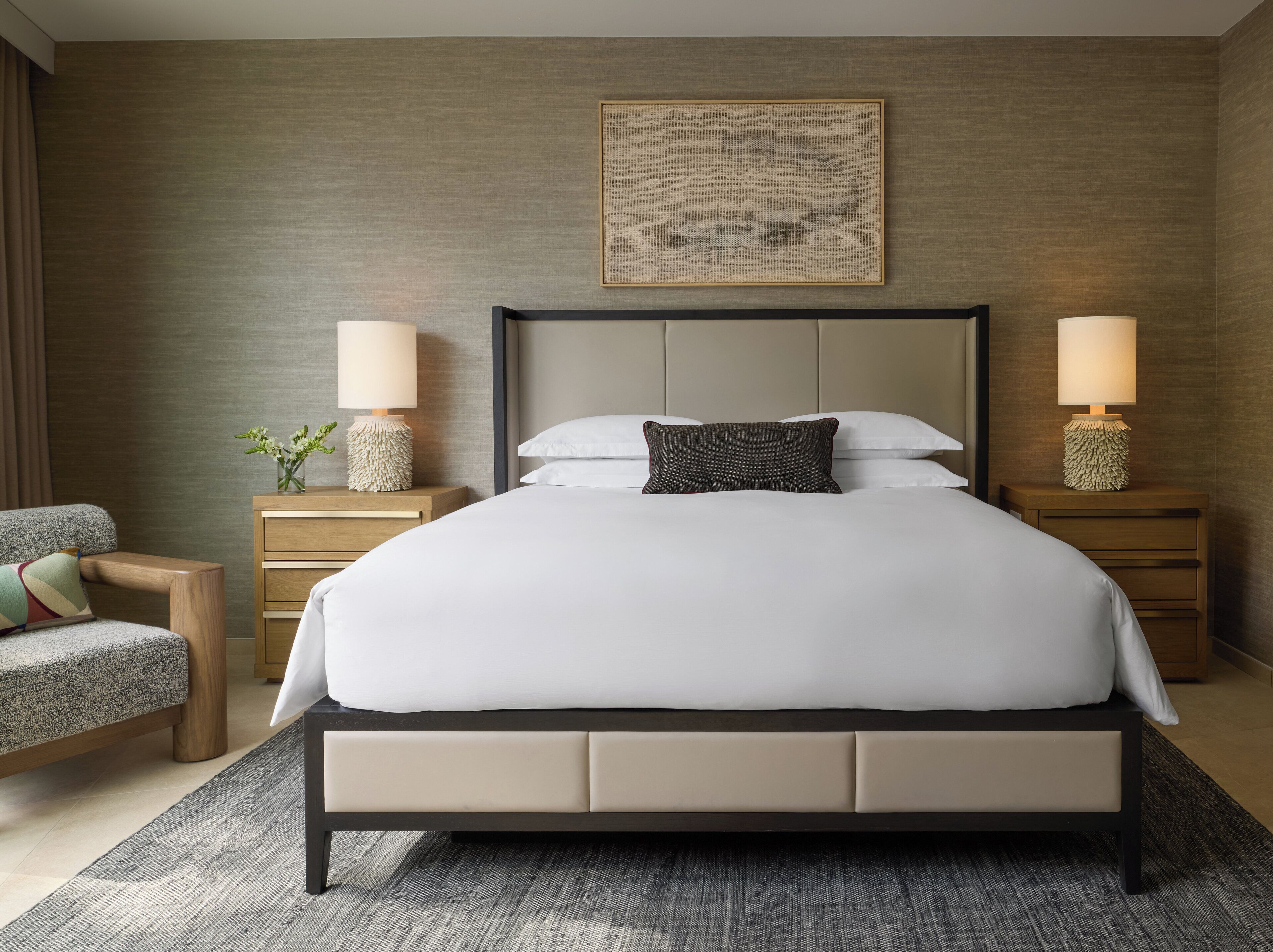 A modern hotel room with a large bed, two bedside tables with lamps, a small armchair, and a framed artwork on the wall.