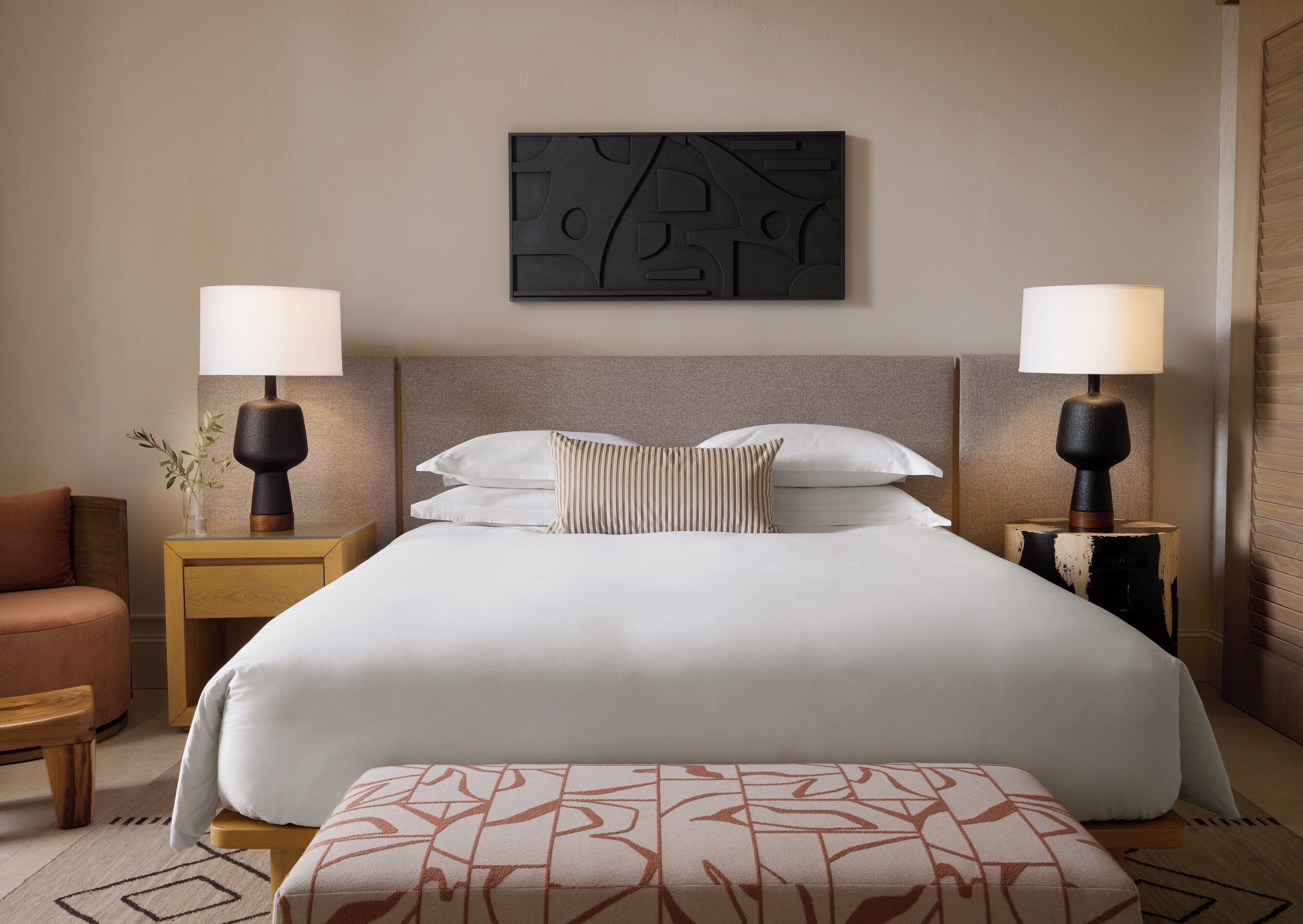 A neatly made bed with white linens, two bedside lamps, a wall-mounted abstract artwork, and a wooden bench with a patterned cushion.