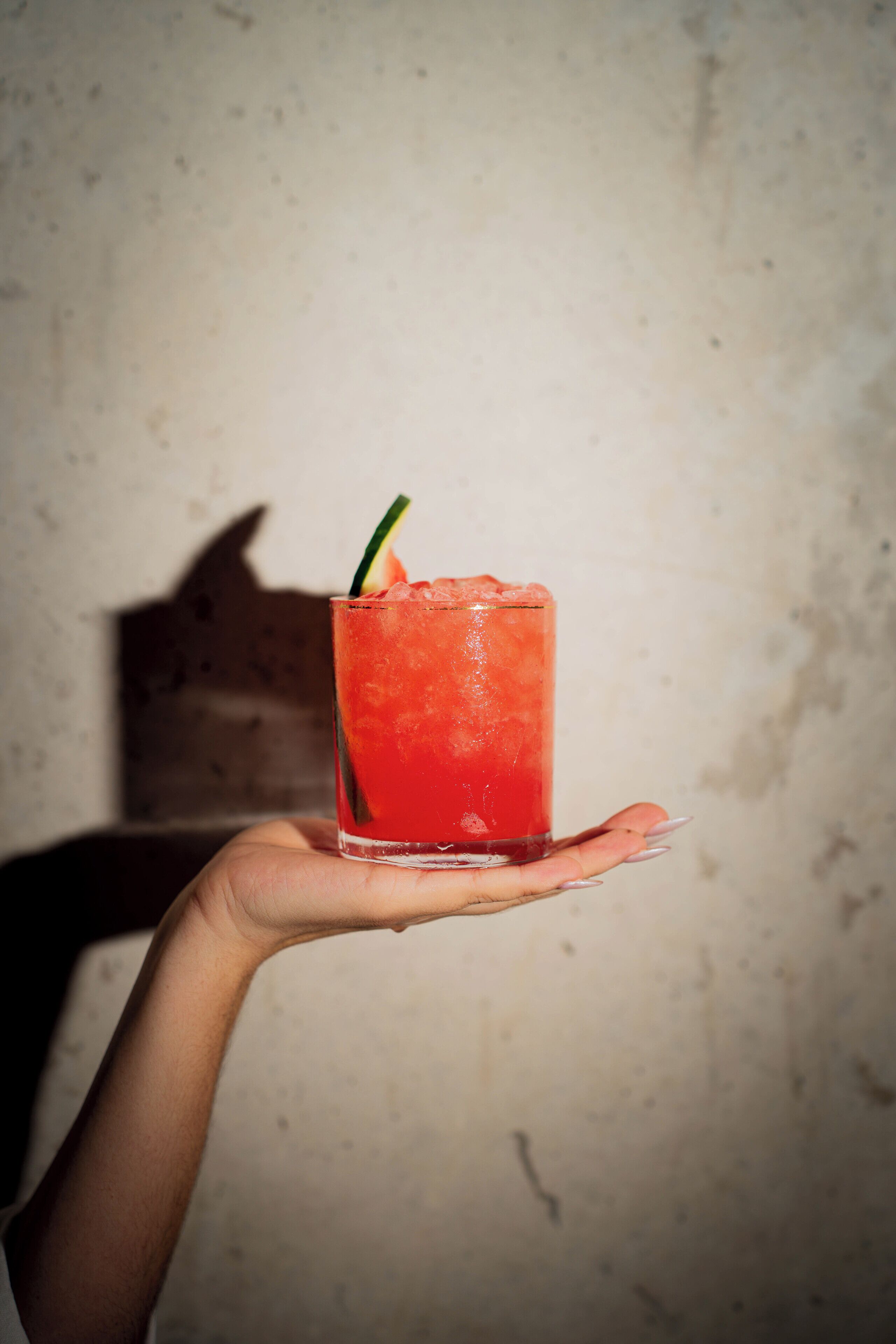 A hand holding a tall glass with a red beverage, possibly a cocktail, garnished with a slice of lime.