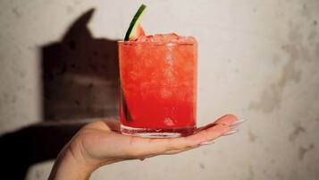 A hand holding a tall glass with a red beverage, possibly a cocktail, garnished with a slice of lime.