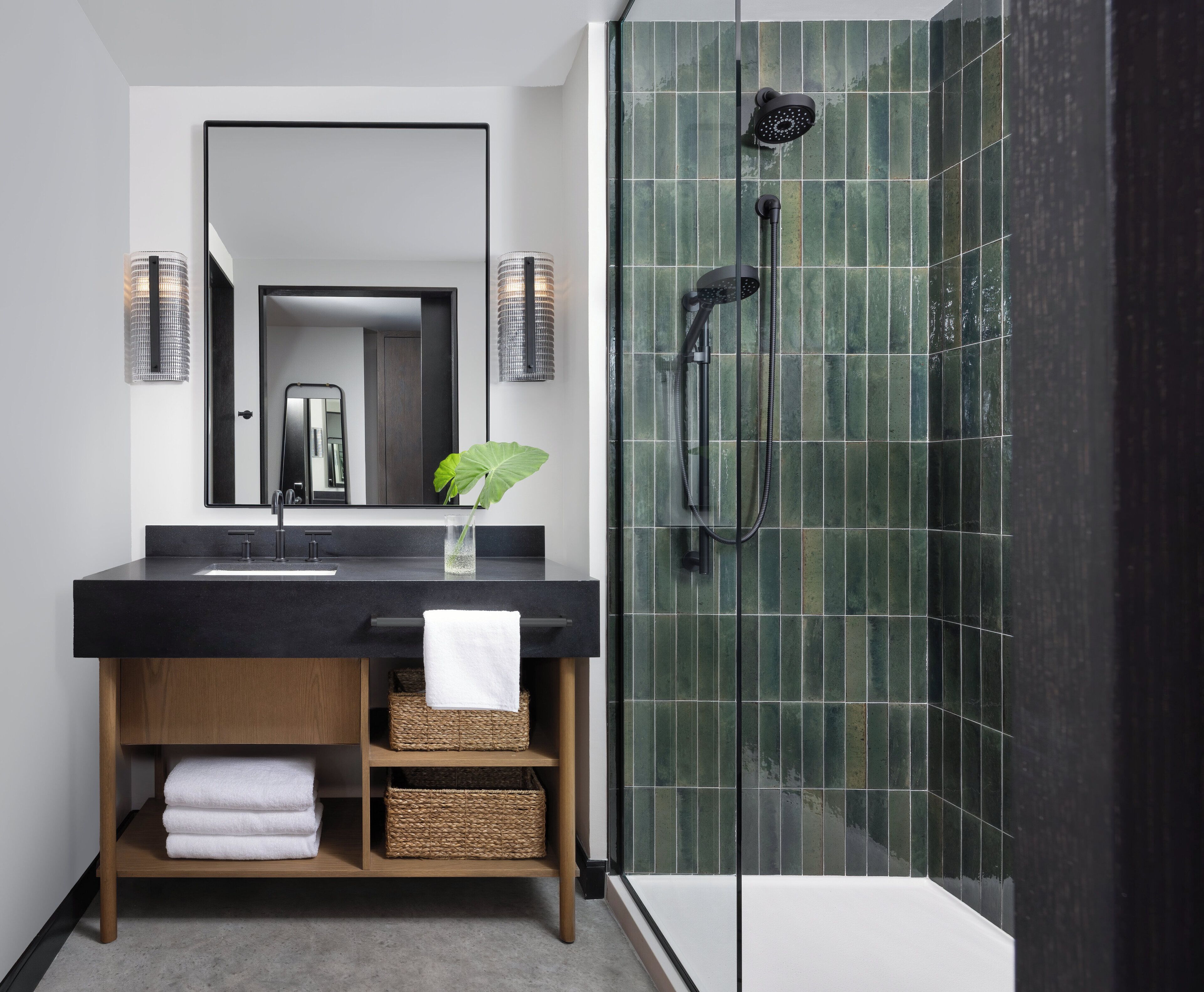 Un bagno moderno con un lavandino nero, uno specchio, una doccia con porta in vetro e una pianta sul lavandino.