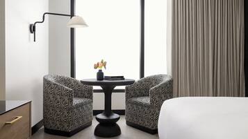 Une chambre d’hôtel avec un lit, deux fauteuils à motifs, une petite table ronde avec un vase, une lampe fixée au mur et une fenêtre avec des rideaux.