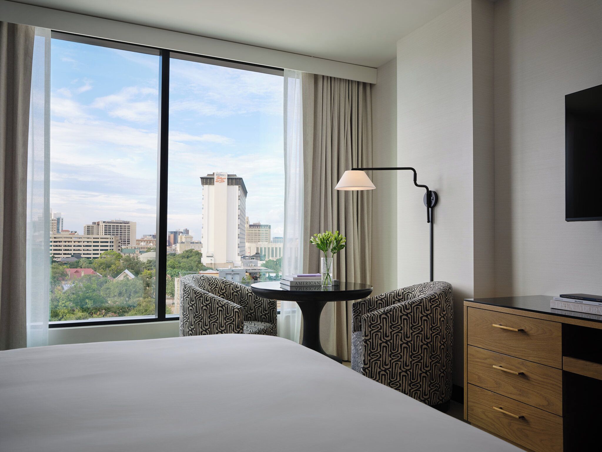 Ein Hotelzimmer mit großem Fenster, einem Bett, einem Schreibtisch mit Lampe, zwei gemusterten Sesseln und einem Fernseher an der Wand.