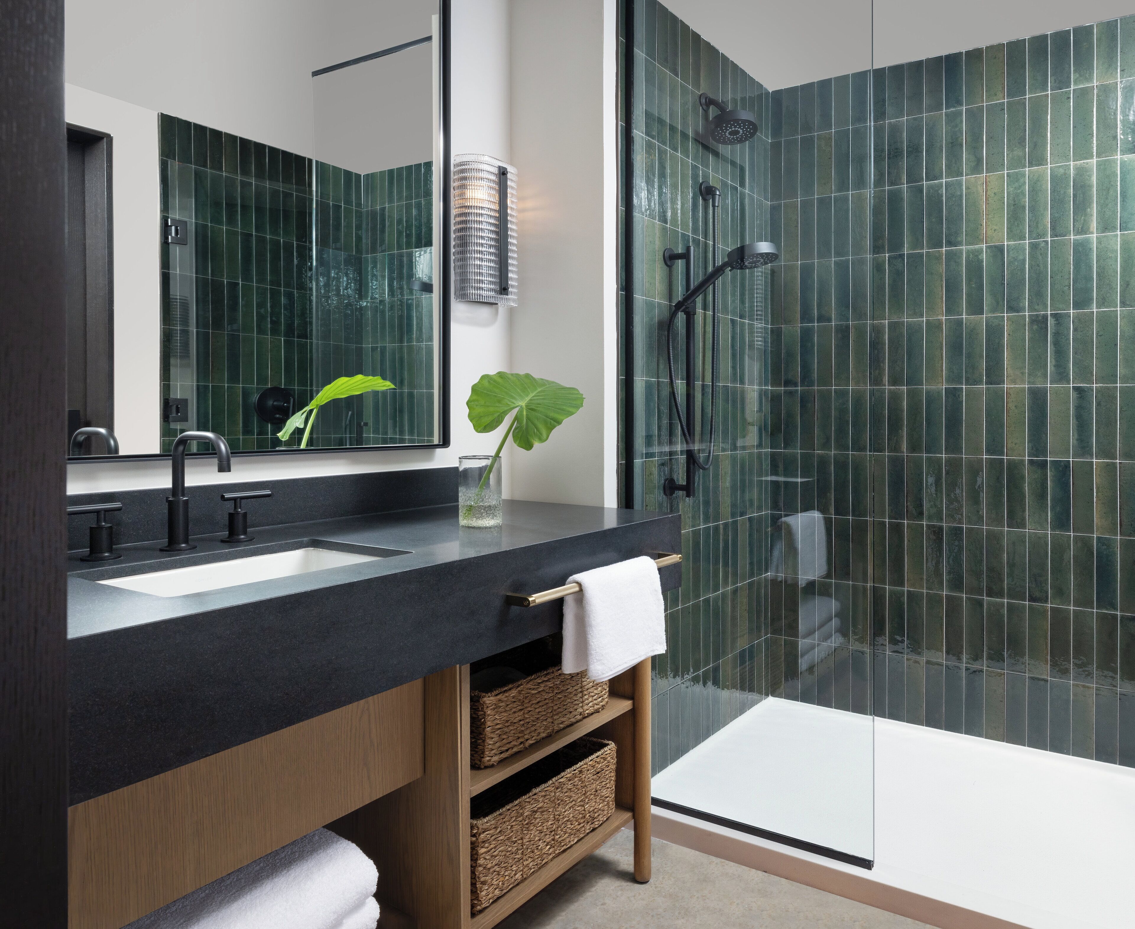 A modern bathroom with a black countertop, two sinks, a large mirror, a shower with a glass door, and a potted plant.