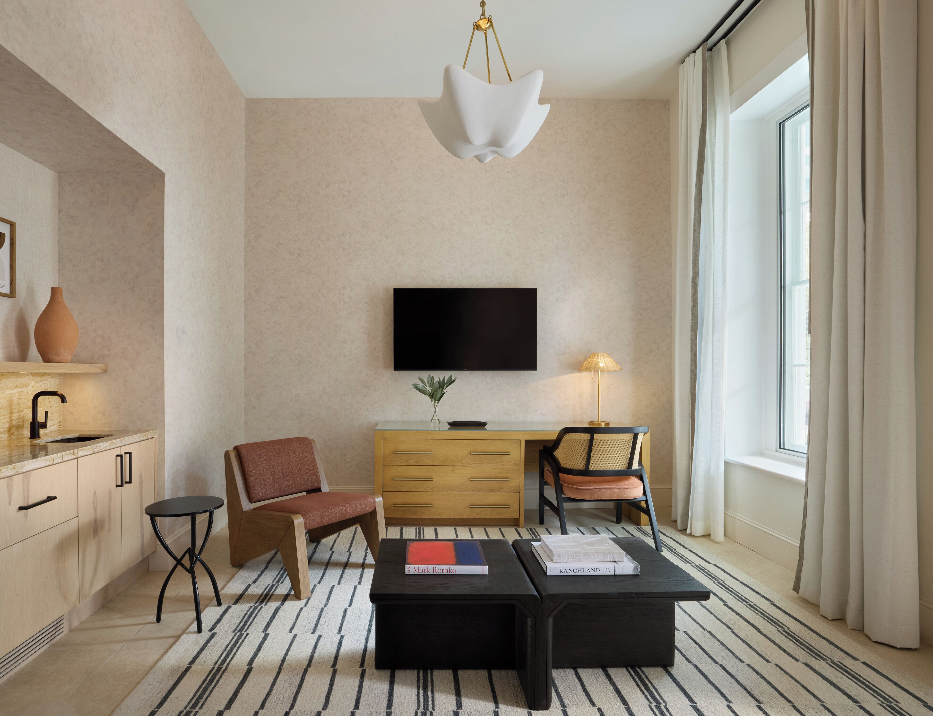 A modern hotel room with a flat-screen TV, a wooden dresser, two chairs, a coffee table, and a large window with curtains.