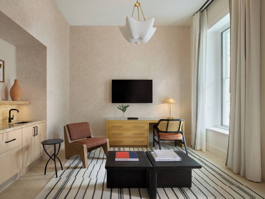 A modern hotel room with a flat-screen TV, a wooden dresser, two chairs, a coffee table, and a large window with curtains.