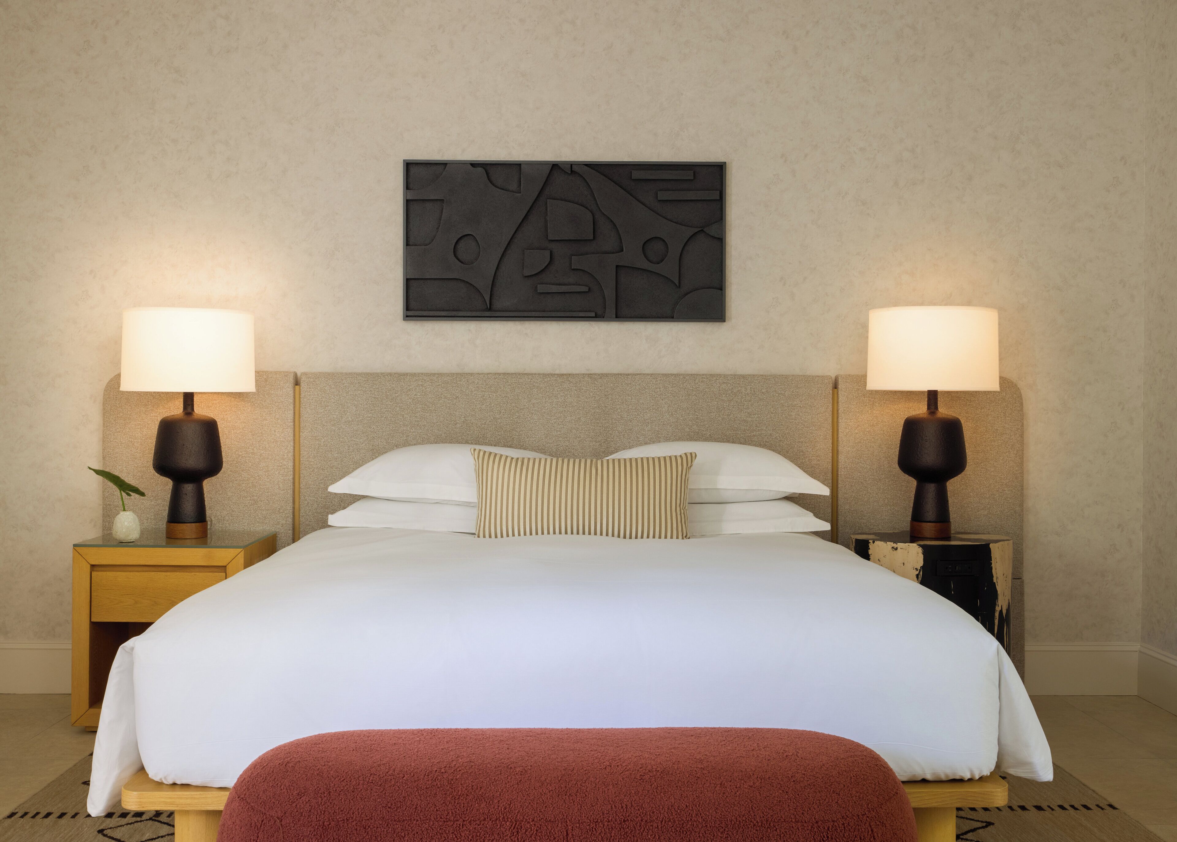 A neatly made bed with white linens, two bedside lamps, and a wall-mounted abstract art piece.