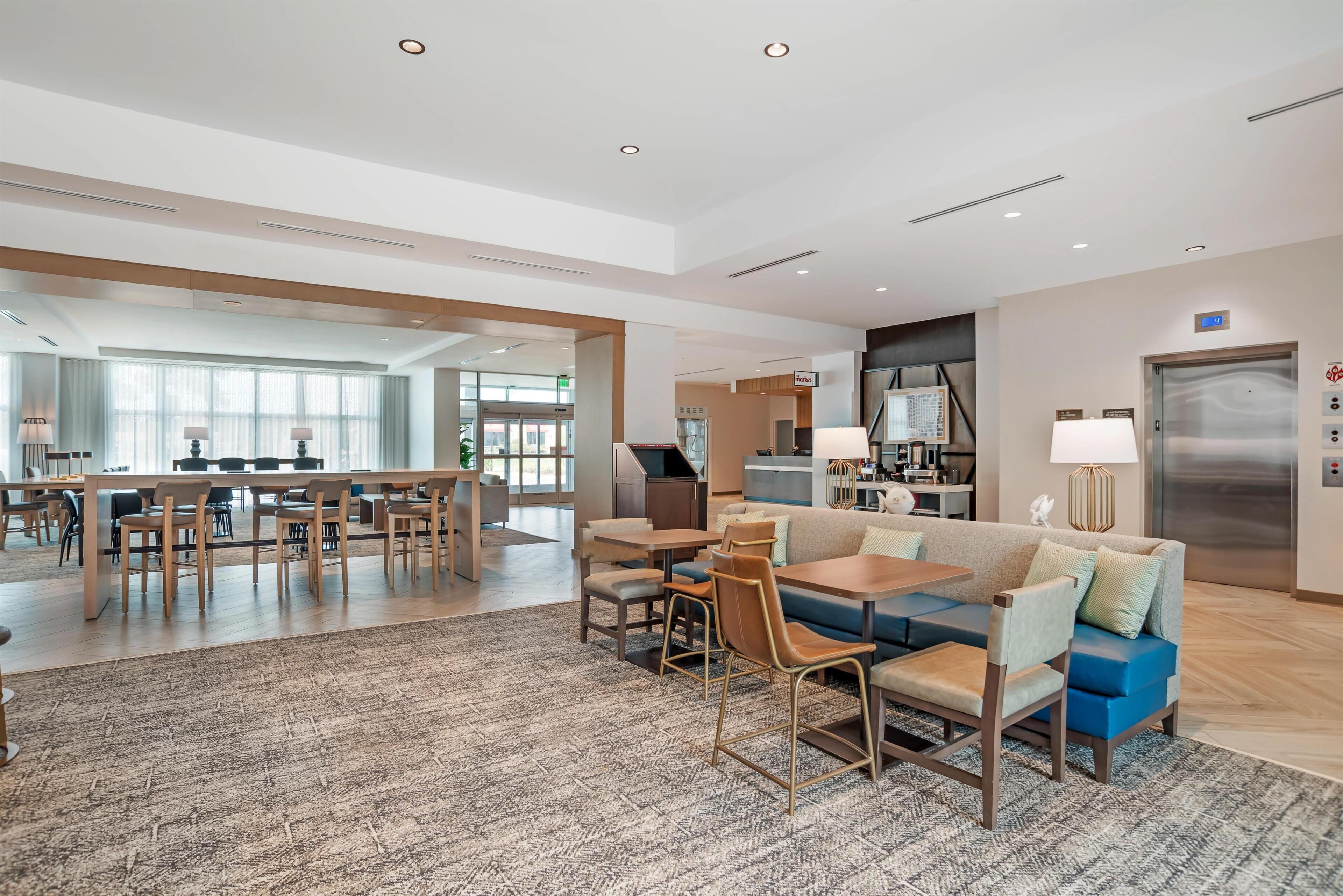 A hotel lobby with a dining area, a sofa, and a kitchen in the background.