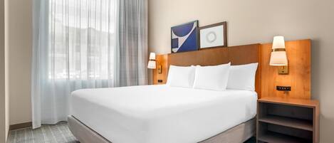 A hotel room with a large bed, two bedside lamps, a nightstand, and a window with sheer curtains.