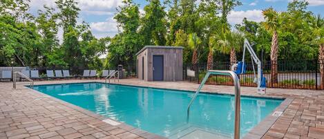 A swimming pool with a small wooden structure, lounge chairs, and a fenced area with palm trees.
