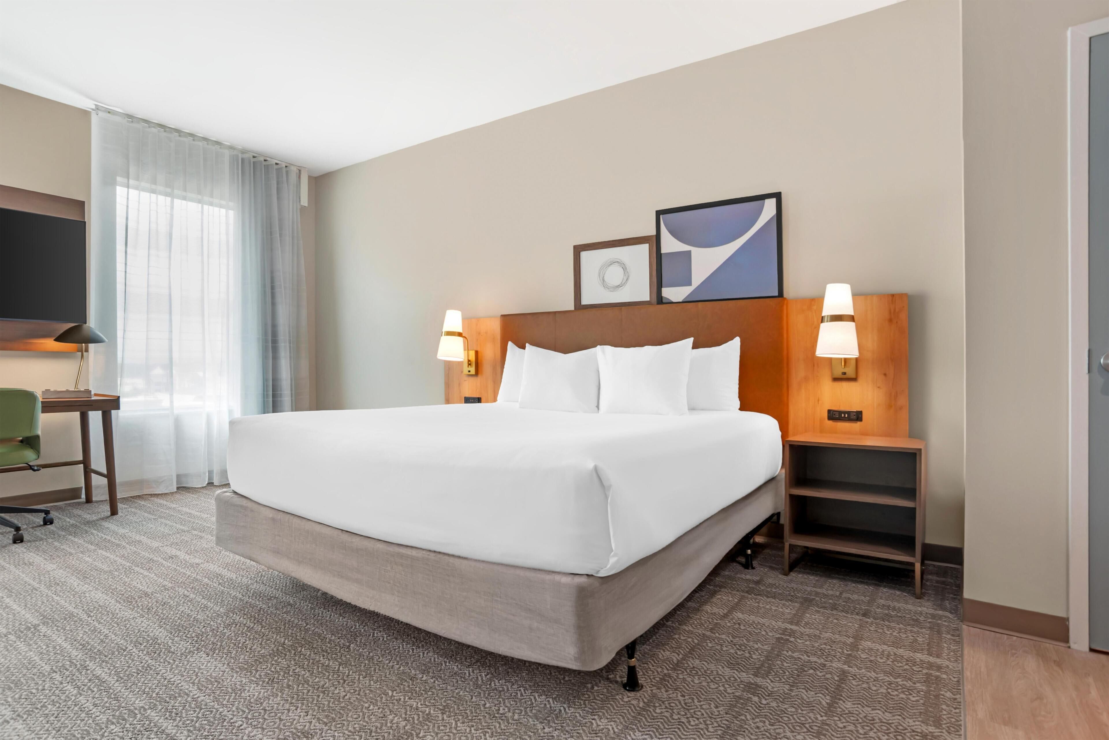A hotel room with a large bed, two bedside tables with lamps, a desk with a chair, and a TV on the wall.