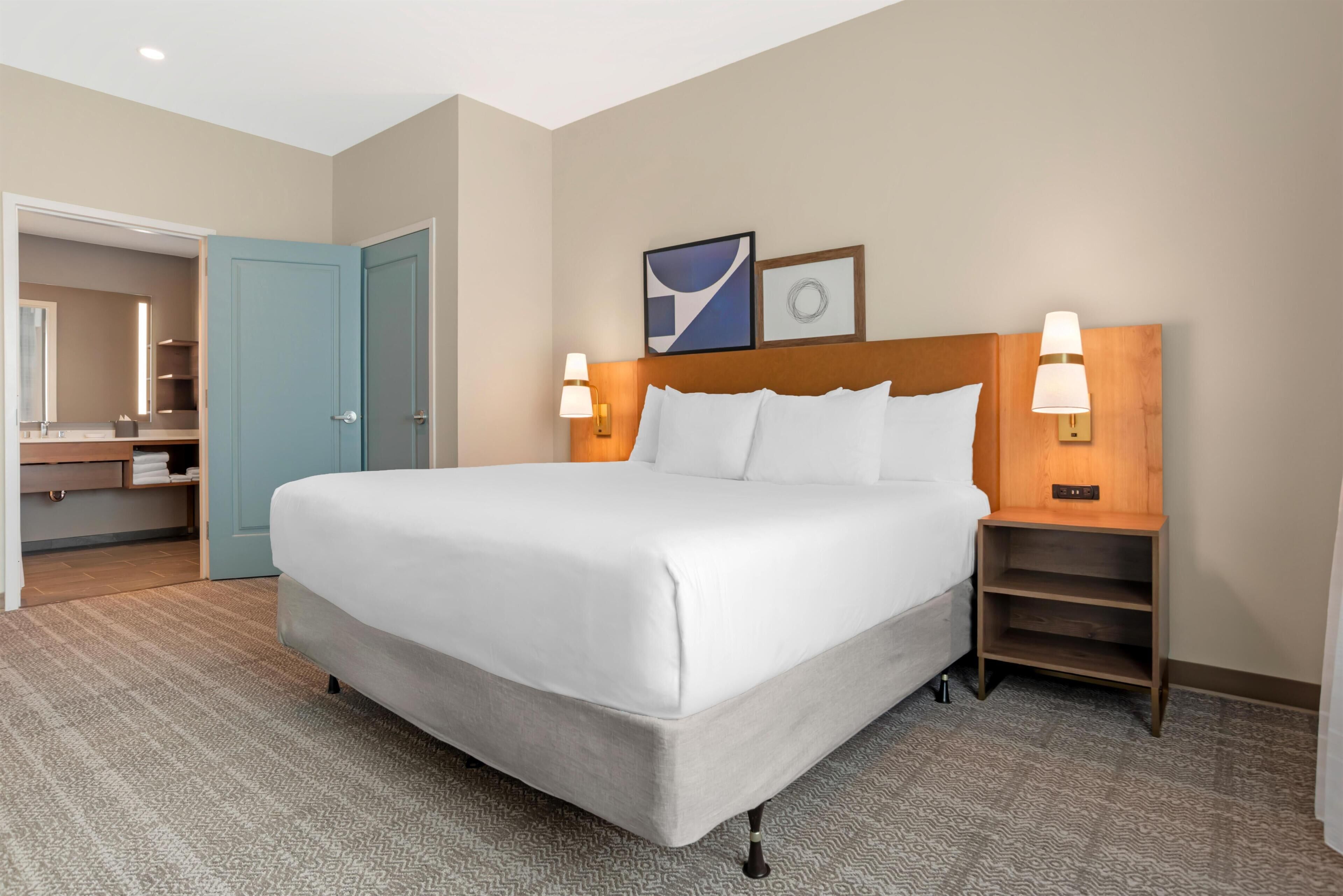 A hotel room with a large bed, two bedside lamps, a wooden nightstand, a headboard with a light fixture, a mirror, and a door leading to a bathroom.