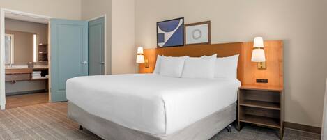 A hotel room with a large bed, two bedside lamps, a wooden nightstand, a headboard with a light fixture, a mirror, and a door leading to a bathroom.