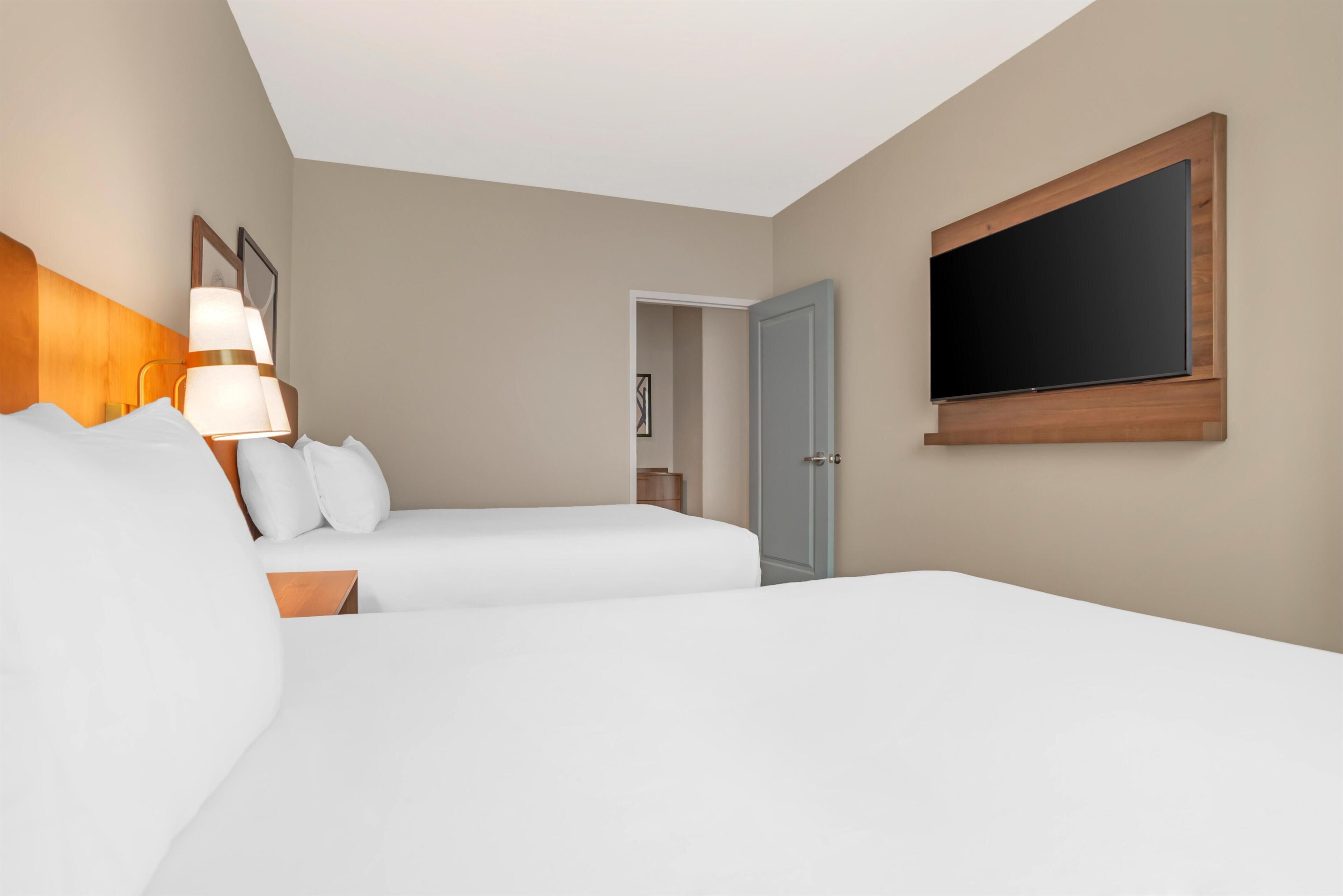 Une chambre d’hôtel avec deux lits, un téléviseur à écran plat fixé au mur et une porte donnant sur une autre pièce.
