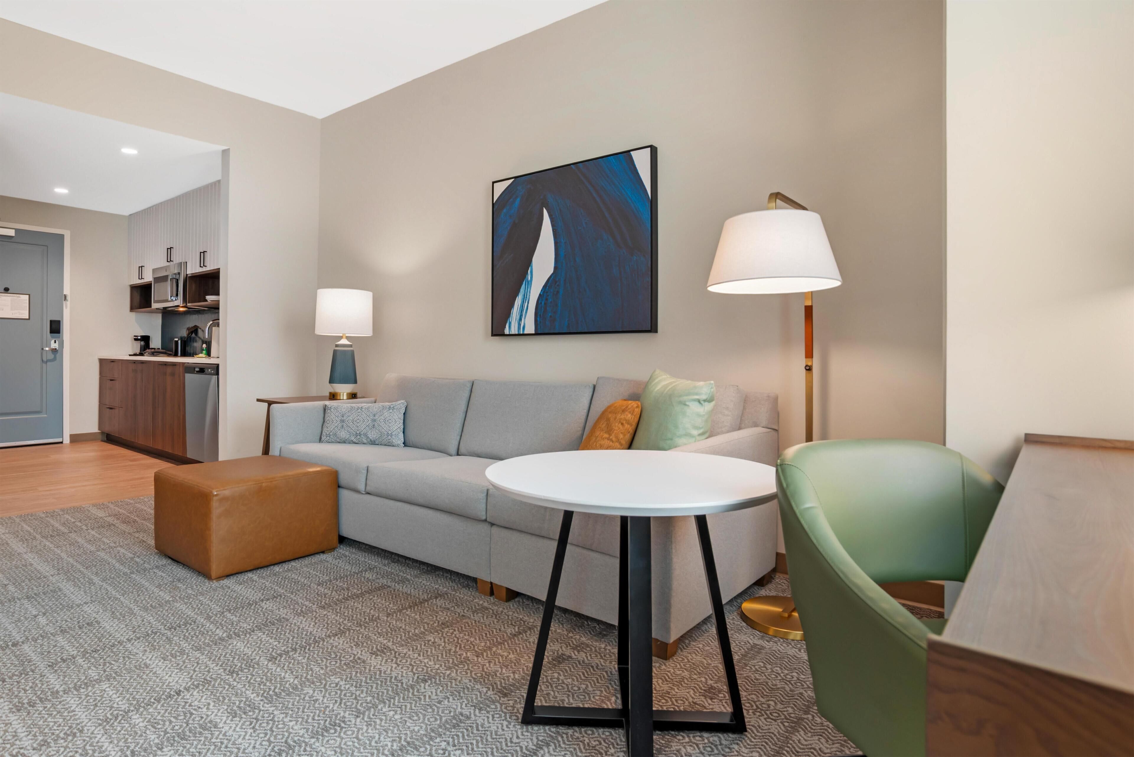 Un salon moderne avec un canapé, un pouf et une table ronde. On y trouve également un coin cuisine avec des placards et un four à micro-ondes. Un tableau est accroché au mur, et une lampe est posée sur une table d’appoint.