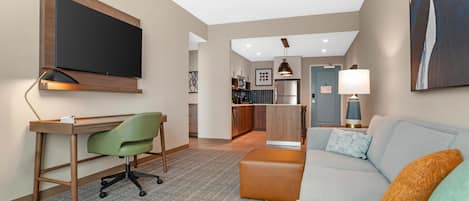 A modern living room with a flat-screen TV, a desk with a chair, a sofa with cushions, a kitchen area, and a dining area.