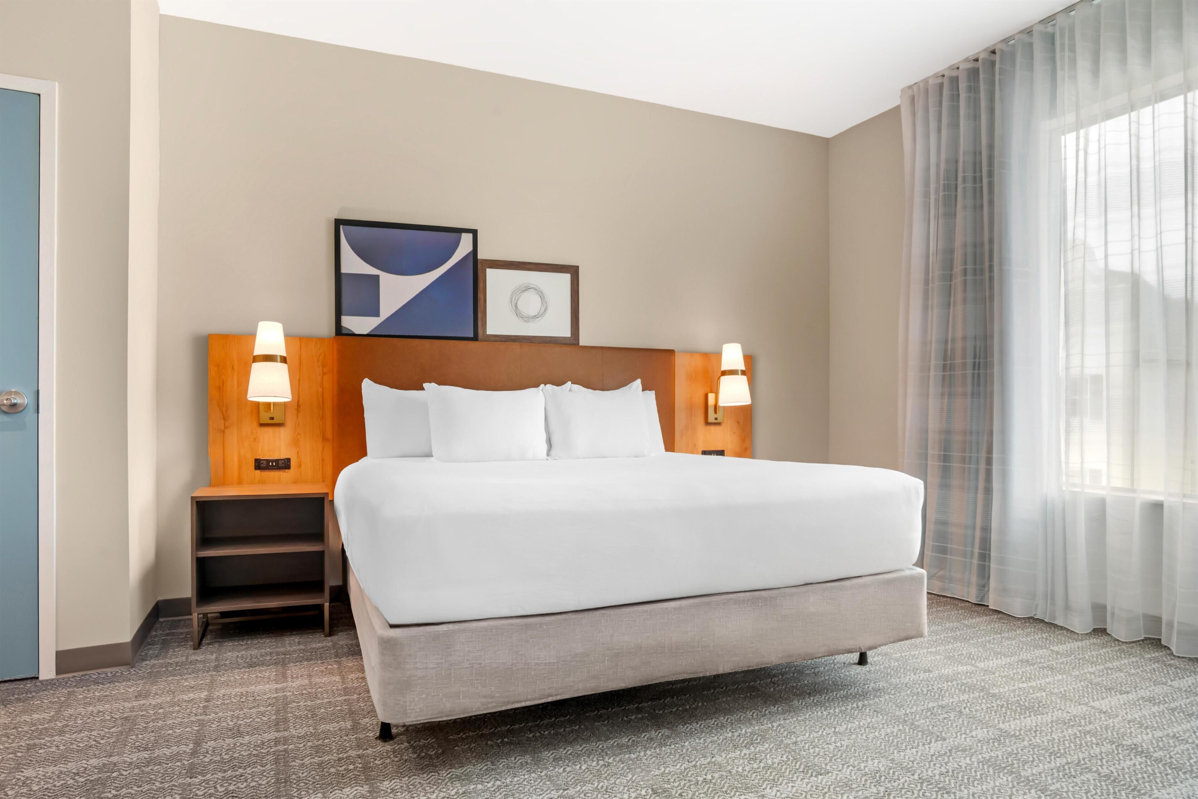 Hotelkamer met een groot bed, twee bedlampjes, een nachtkastje, een lijst met een afbeelding en een raam met dunne gordijnen.