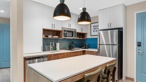 A modern kitchen with white cabinets, a stainless steel refrigerator, a microwave, and a large island with bar stools. - Drury Inn & Suites Destin Miramar Beach (Miramar Beach)