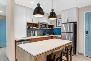 A modern kitchen with white cabinets, a stainless steel refrigerator, a microwave, and a large island with bar stools. - Drury Inn & Suites Destin Miramar Beach (Miramar Beach)