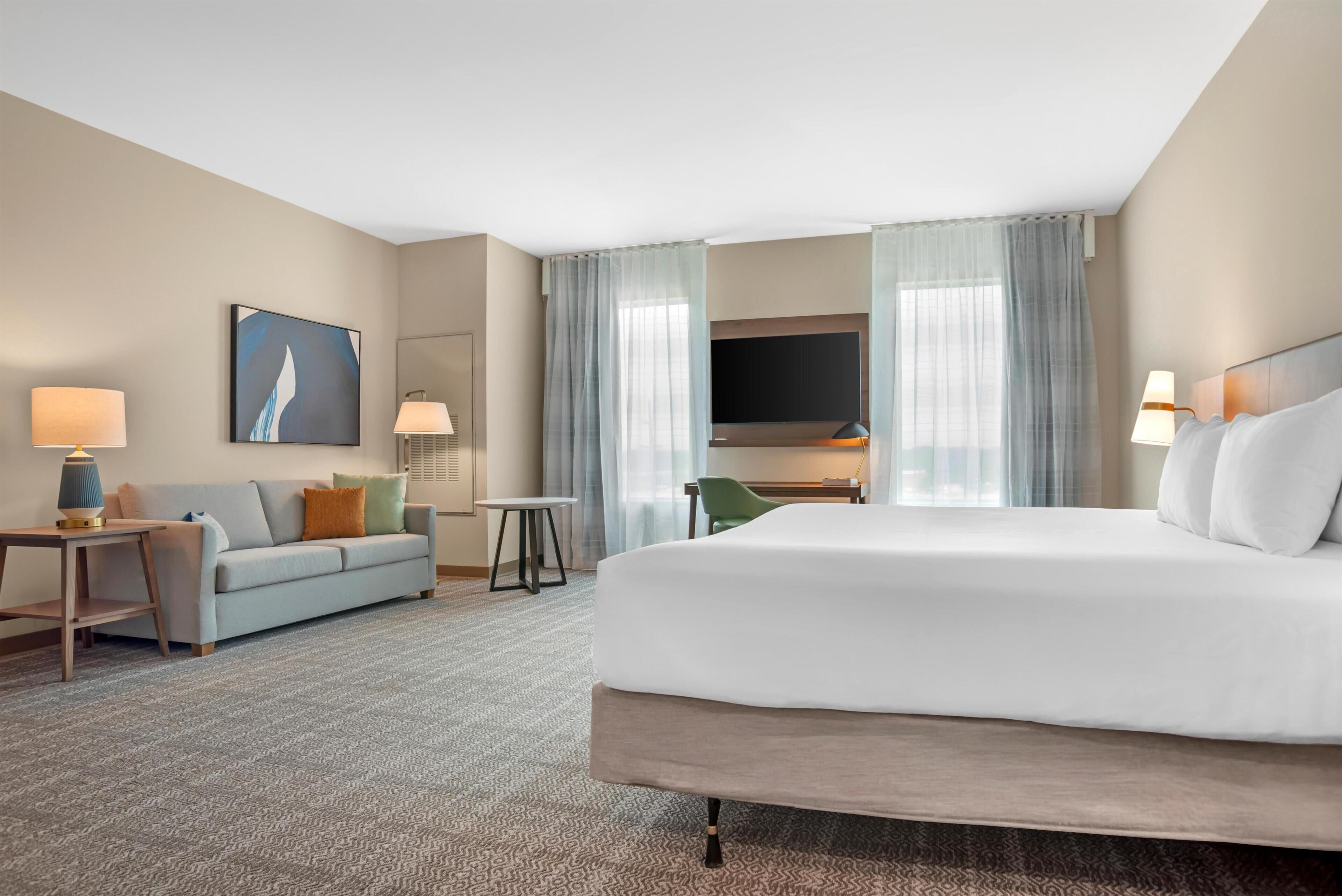 A hotel room with a bed, a sofa, a desk, a TV, and a painting on the wall.