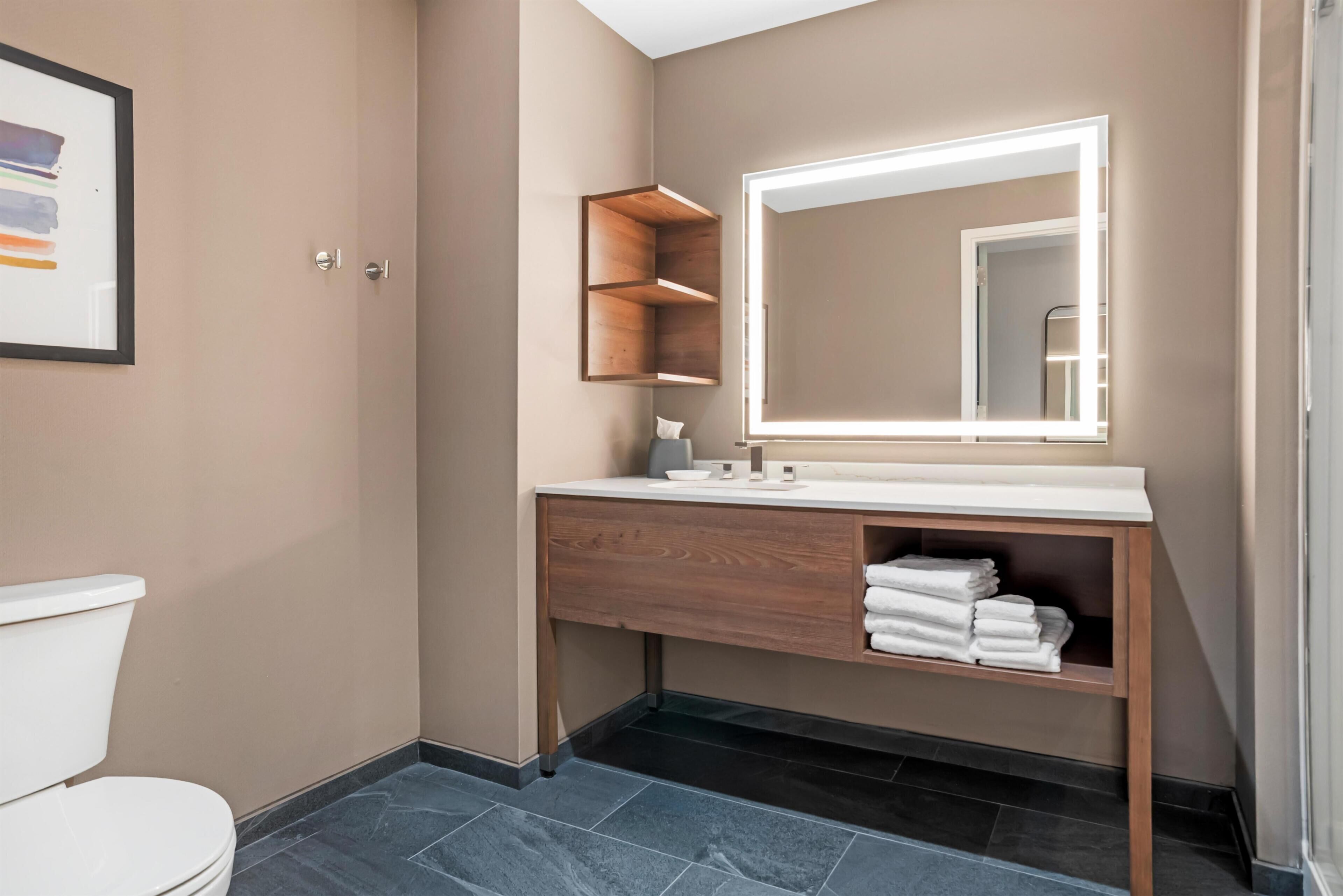 A bathroom with a toilet, a vanity with a mirror, and a shelf with towels.