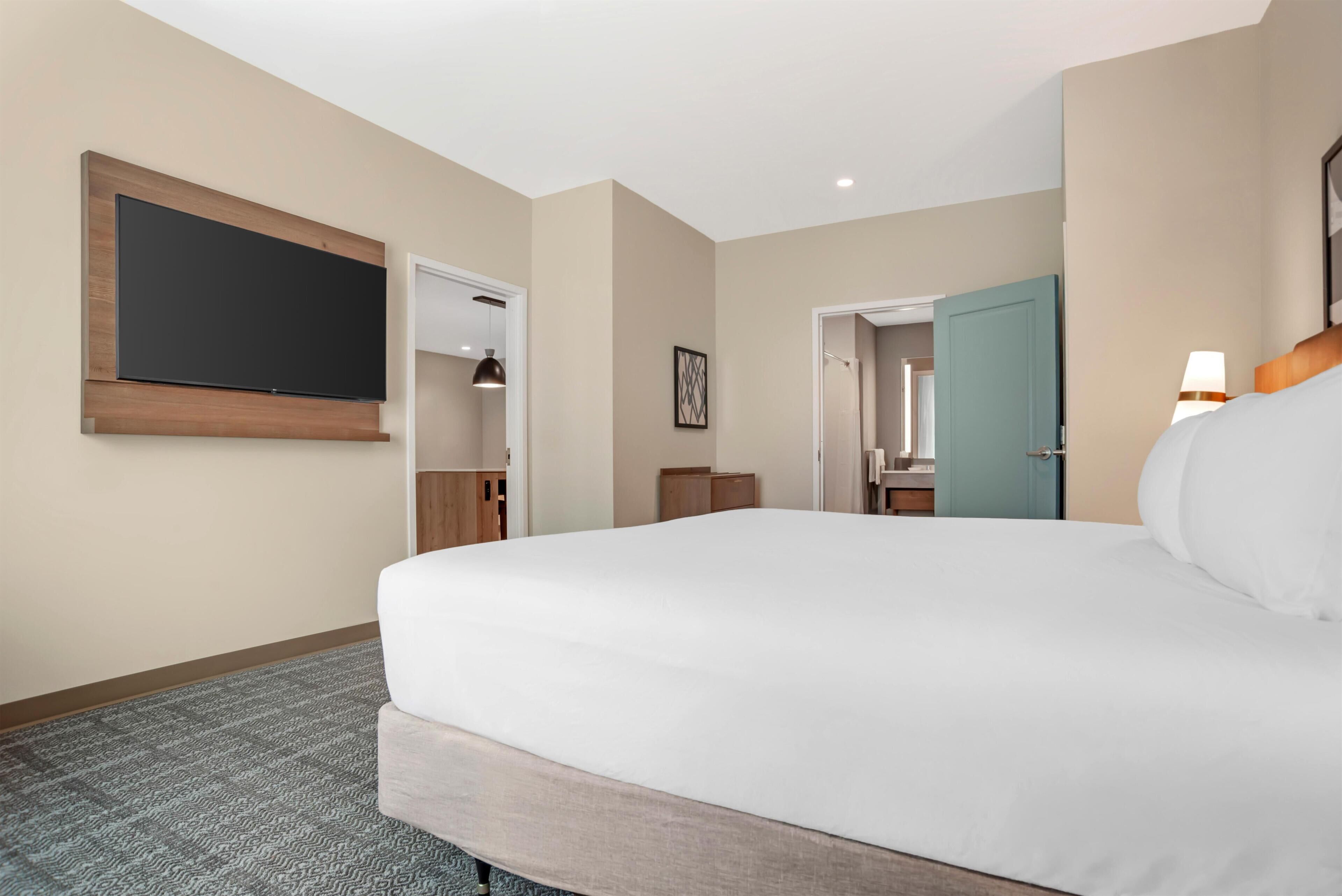 Ein Hotelzimmer mit einem großen Bett, einem flachbildfernseher an der Wand, einer Tür zu einem Badezimmer und einem kleinen Tisch mit einer Lampe.