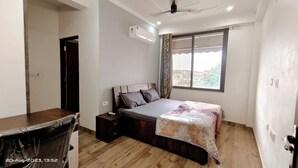 A bedroom with a bed, a desk, a chair, a window with curtains, and an air conditioning unit. - OSHO Villa Homestay, 5 minutes to the Jaipur Airport, Garden View AC Rooms,  (Jaipur)